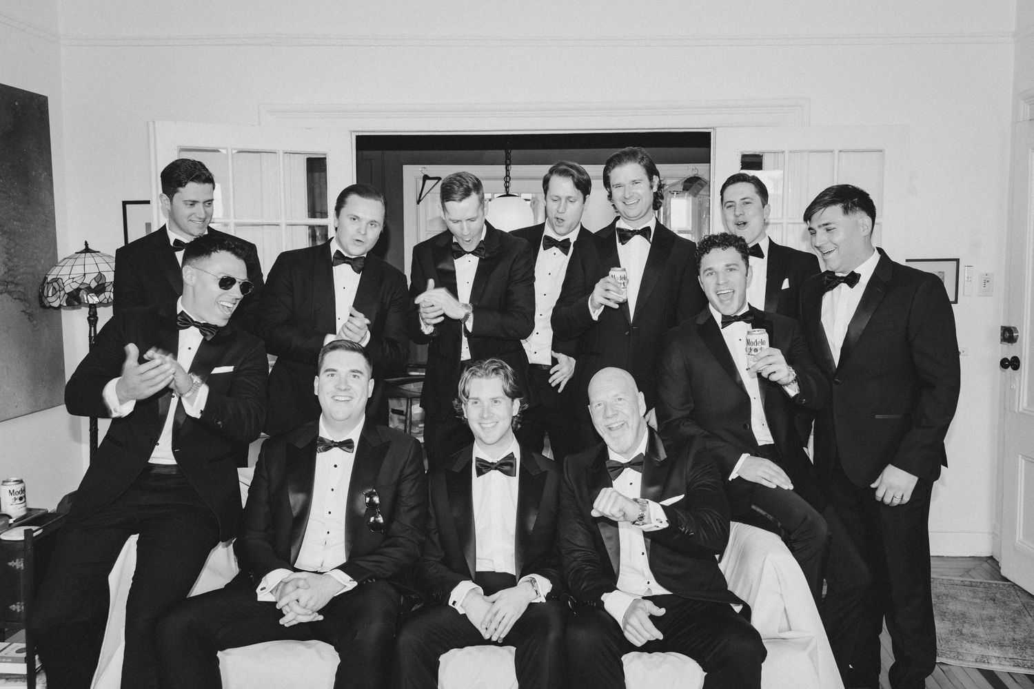 Black and white photo of groomsmen in tuxedos celebrating together, raising glasses in a toast at a wedding.