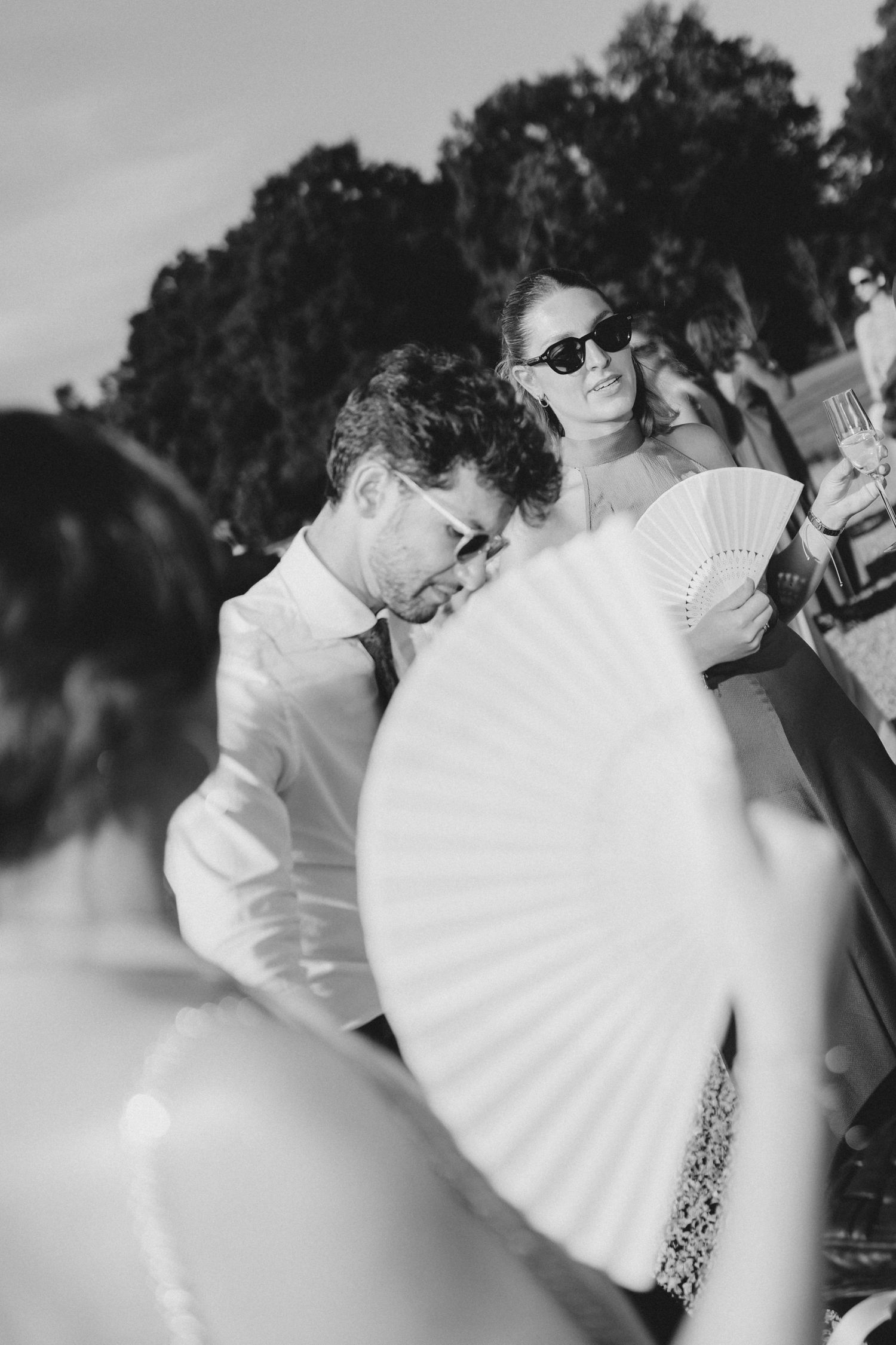Black and white photo of wedding guests using decorative paper fans outdoors during daytime celebration.