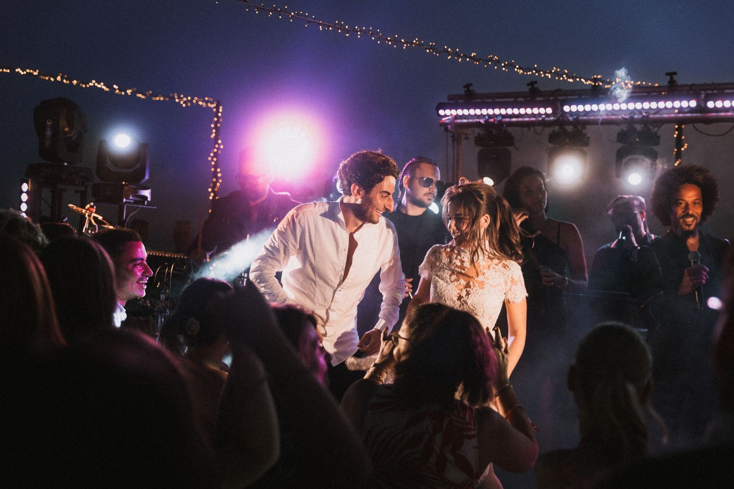 Wedding party dancing under string lights at nighttime reception.