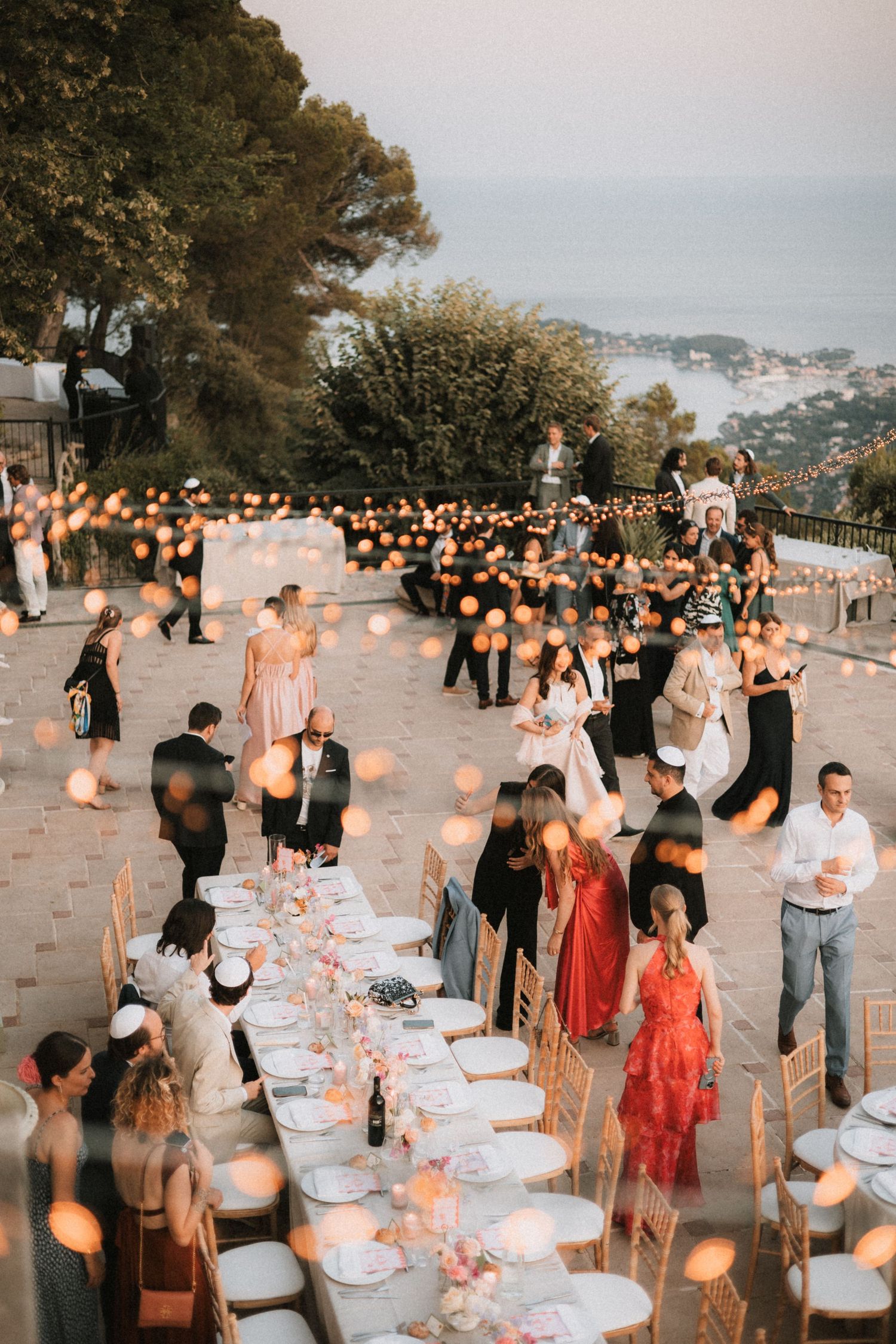Outdoor wedding reception setup with glowing lanterns and tables arranged along a coastal viewpoint.