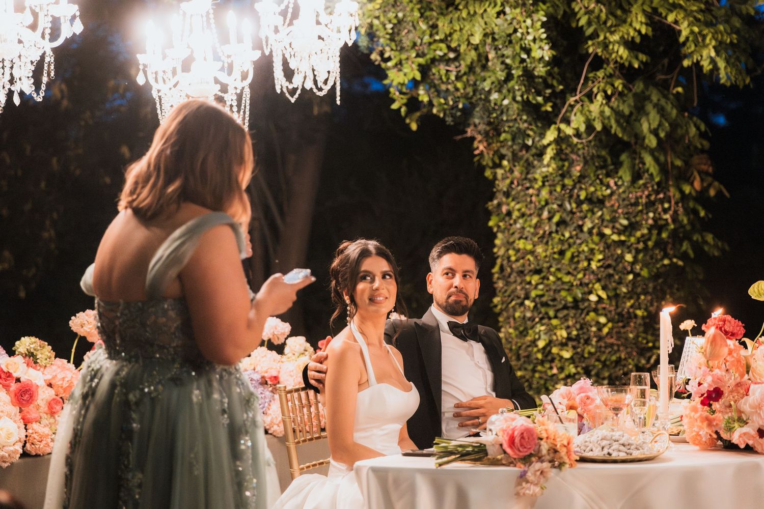 Bride and groom seated at wedding reception table with pink floral arrangements while guest gives speech under chandelier.