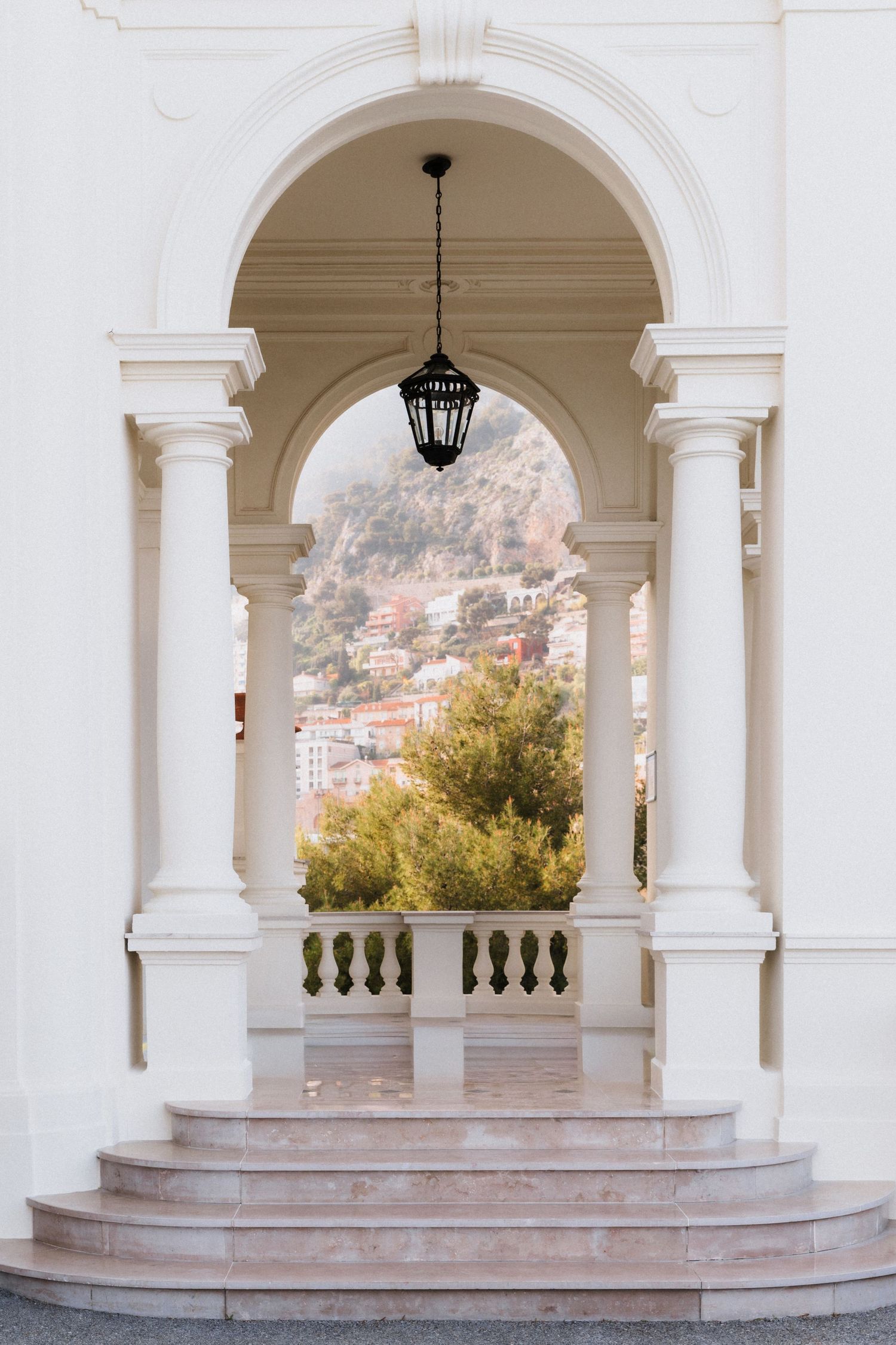 Villa La Vigie, Monaco - Elegant white arched colonnade with hanging lantern, marble steps, and scenic hillside village view beyond.