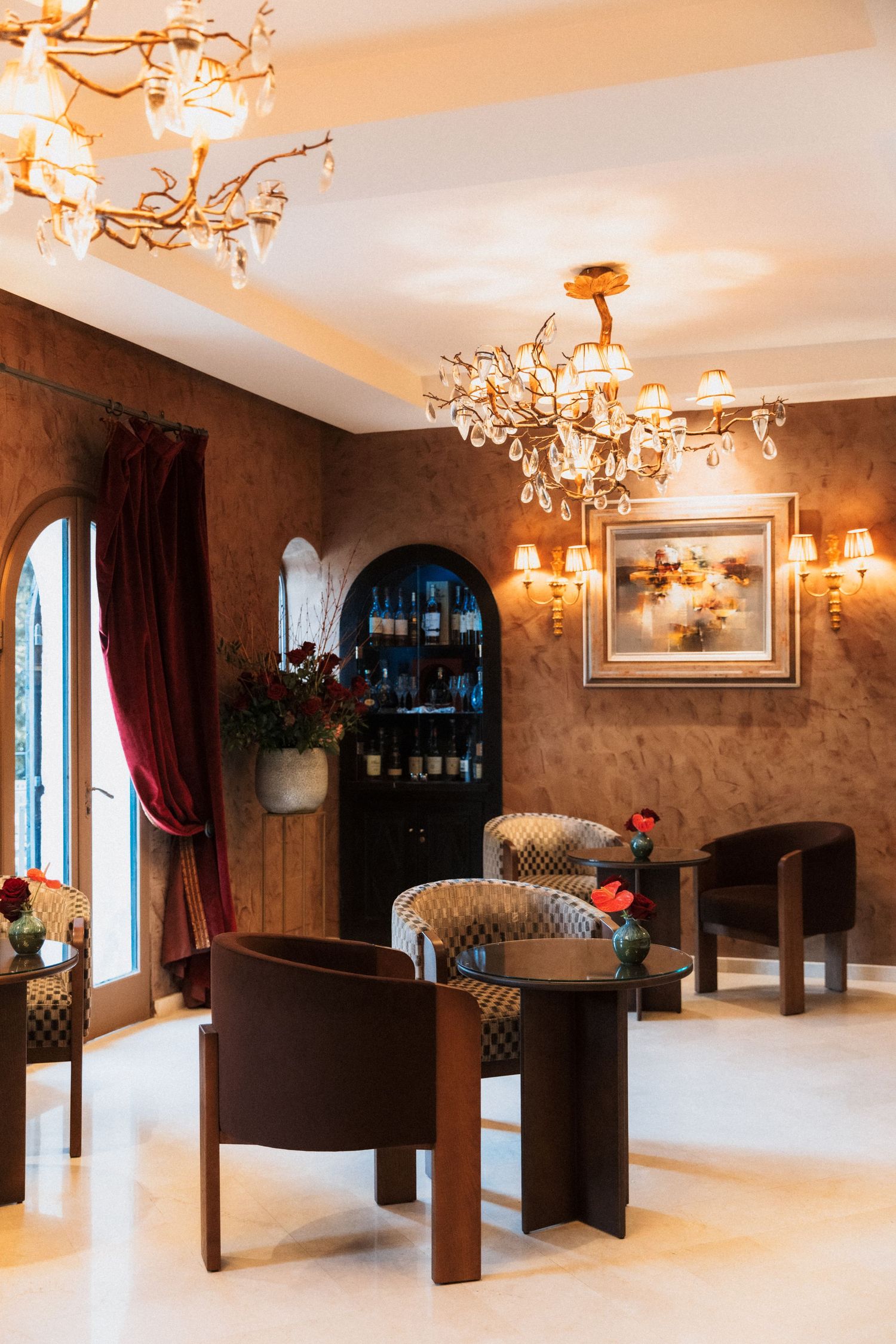 Château Eza - Elegant hotel lounge with ornate chandeliers, brown leather chairs, arched wine cabinet, and warm wall sconces.