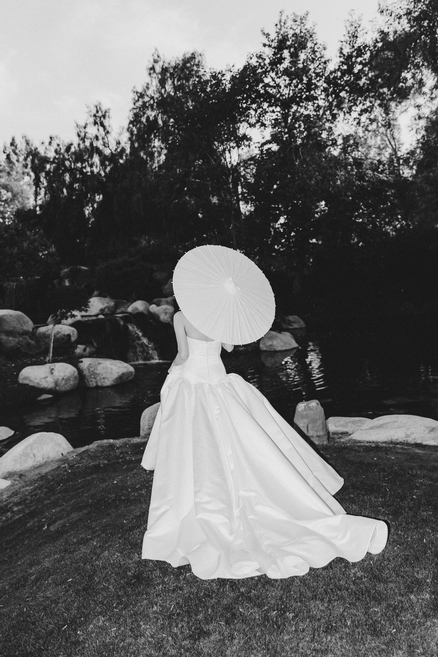 A black and white photo of a bride in a flowing white dress holding a parasol while standing on grass near rocks.