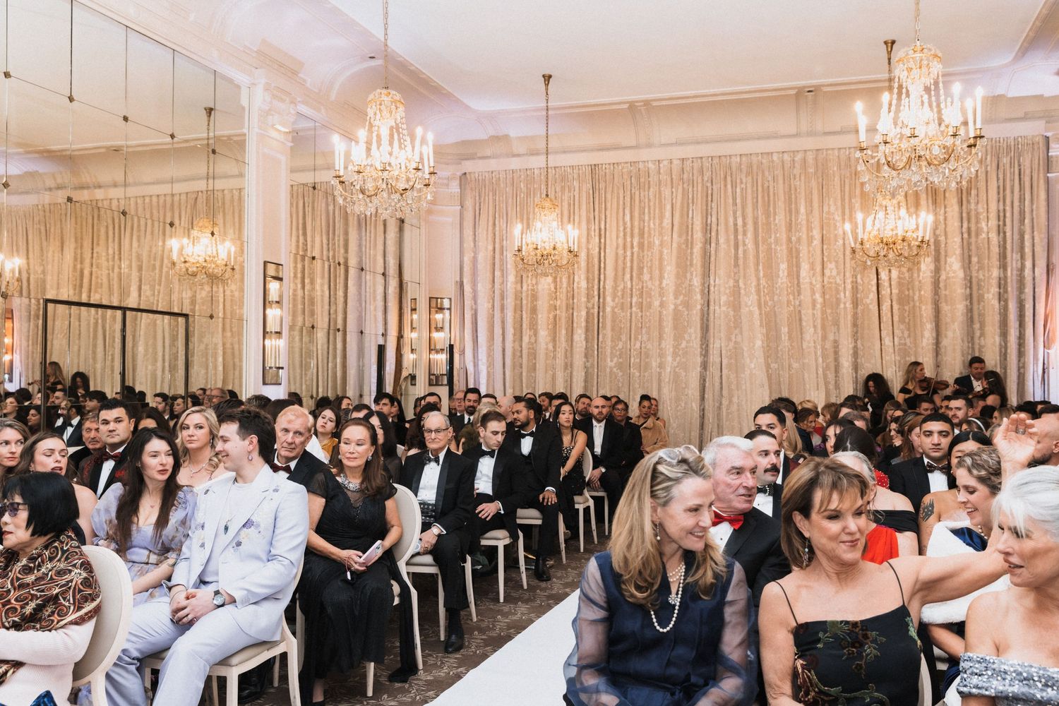Wedding guests watch attentively during an elegant ballroom ceremony with crystal chandeliers and white decor.