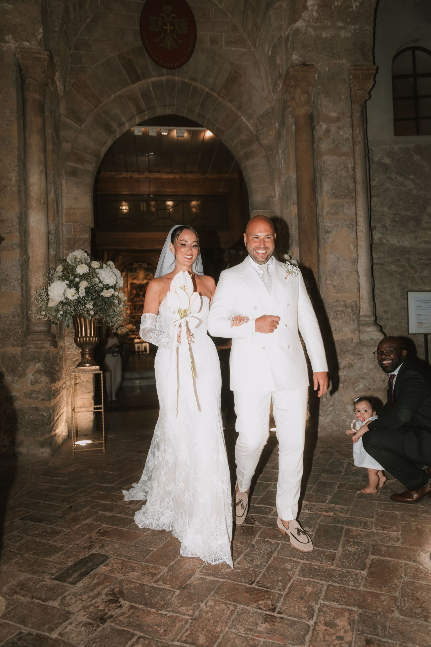 Bride and groom in white attire exit stone chapel during elegant wedding ceremony with floral arrangements.