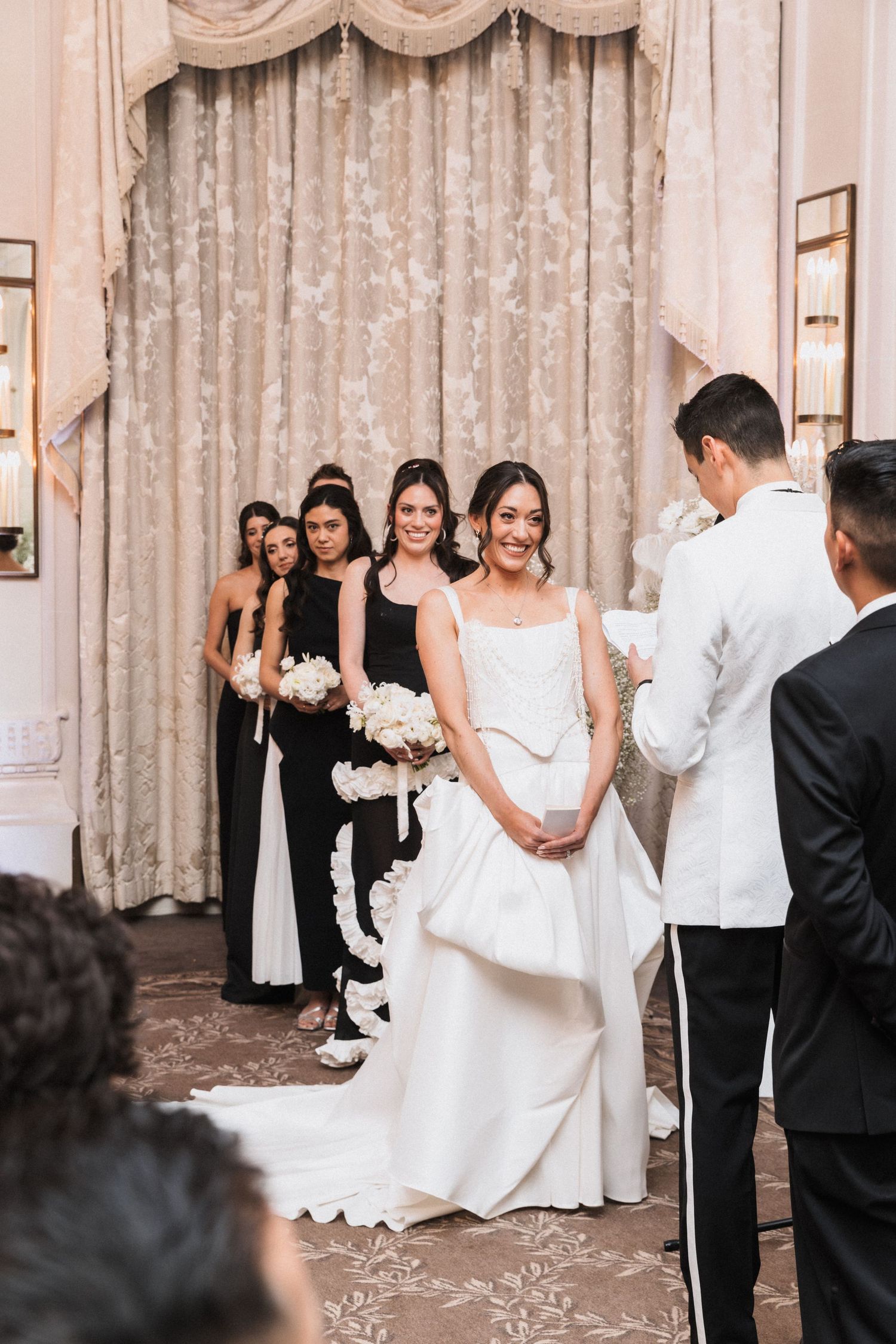 A formal wedding ceremony in progress in an elegant ballroom with lace curtains and classical decor.