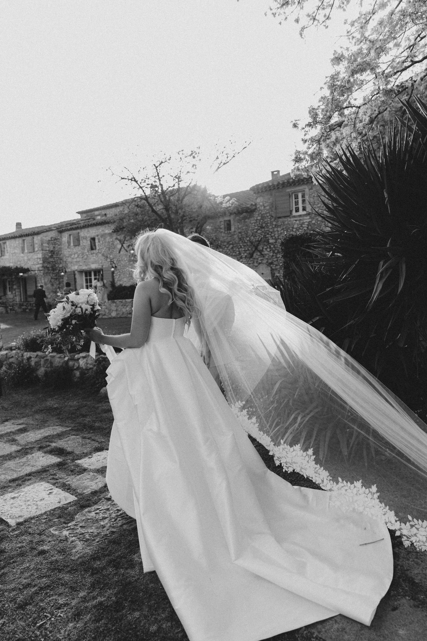 A flowing white wedding veil billows dramatically in the wind against a garden backdrop.