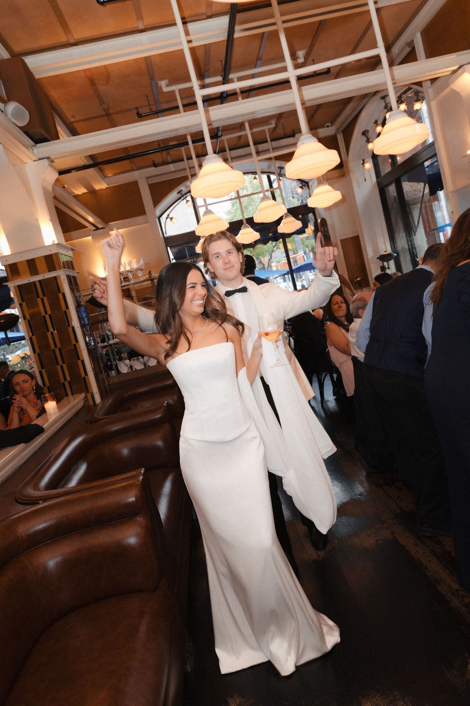 Bride and groom in white attire dancing joyfully among guests at elegant indoor wedding reception.