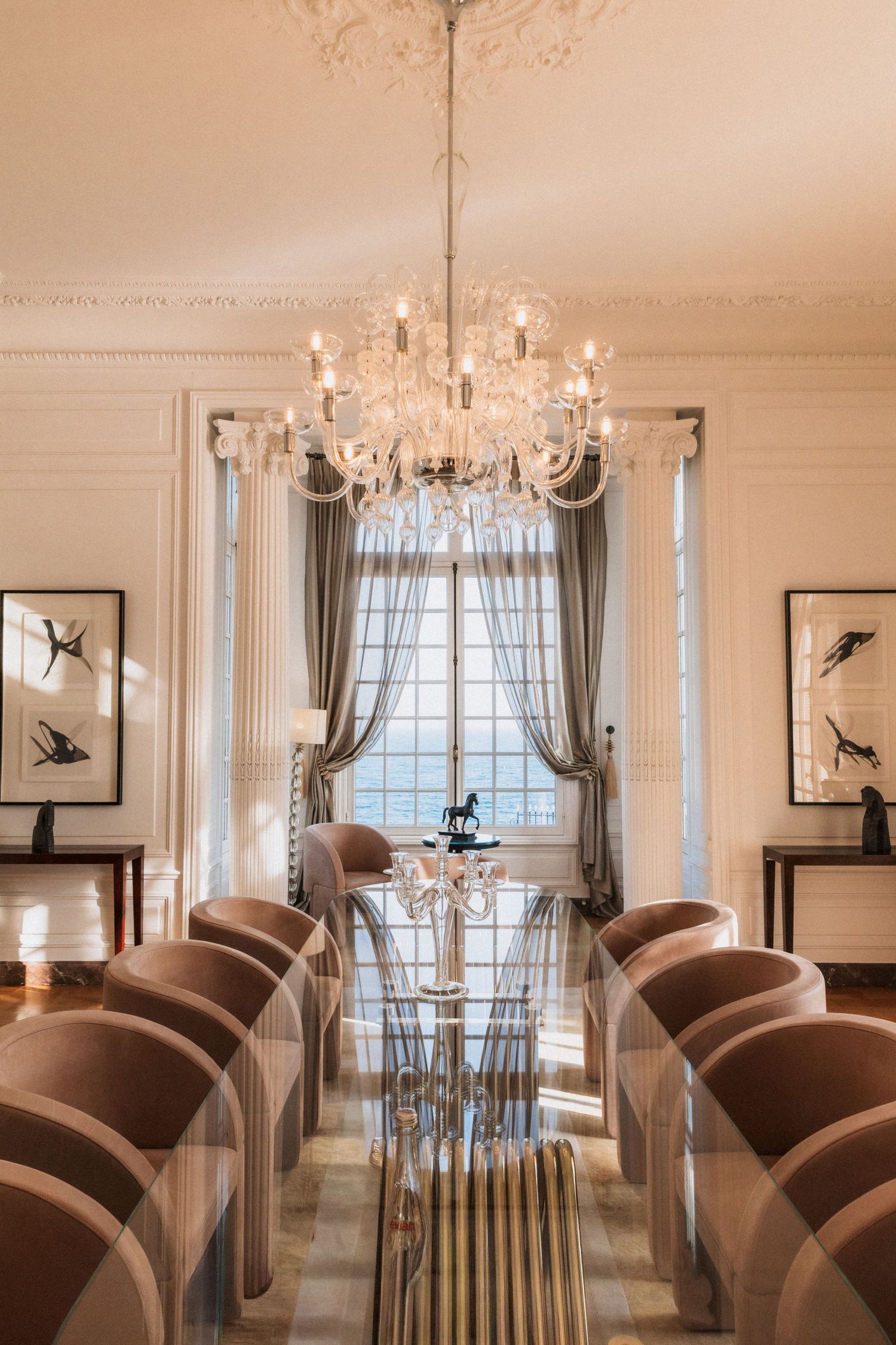 Villa La Vigie, Monaco - Elegant dining room with crystal chandelier, glass table, velvet chairs, tall windows, and ornate molded ceiling.