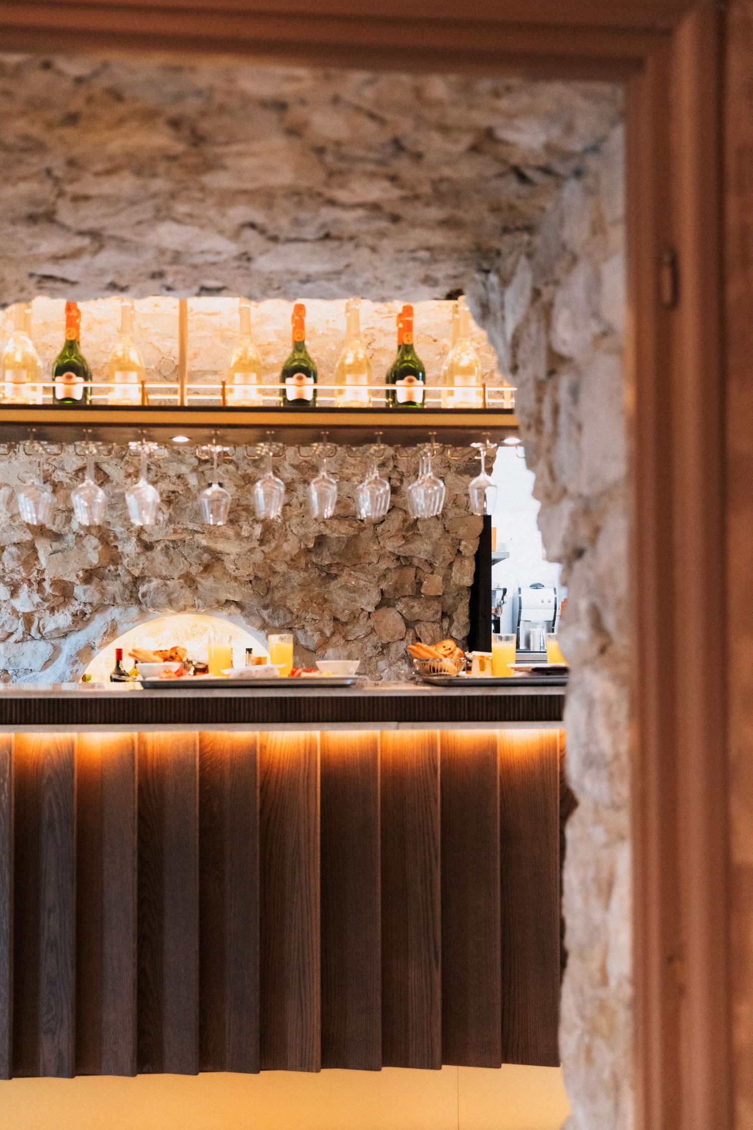 Château Eza - Elegant bar with stone walls, illuminated wine bottles on shelves, hanging glasses, and warm wooden counter.