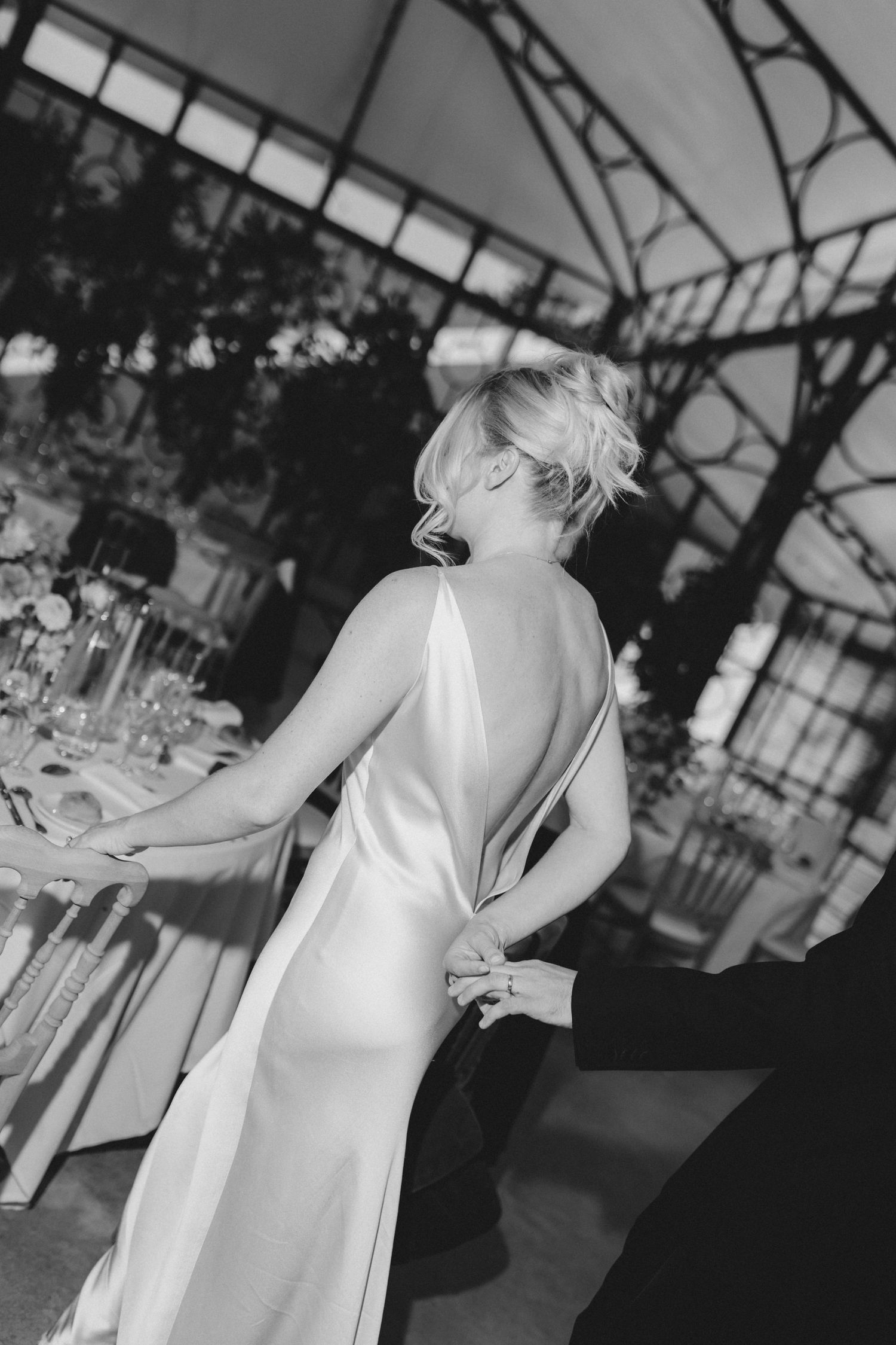 A bride in an elegant silk slip dress laughs joyfully during her wedding reception in a glass-ceiling venue.
