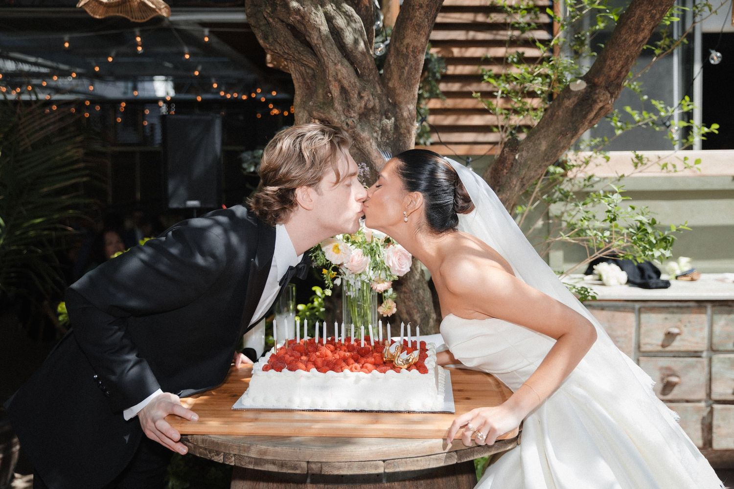 Bride and groom sharing a kiss over a white wedding cake with strawberries and candles at an outdoor reception.