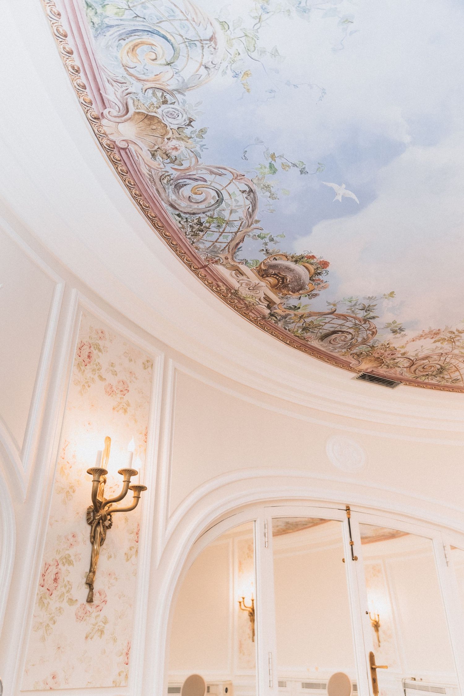 Ornate baroque ceiling with delicate painted details and gold trim, featuring a crystal wall sconce in an elegant room.