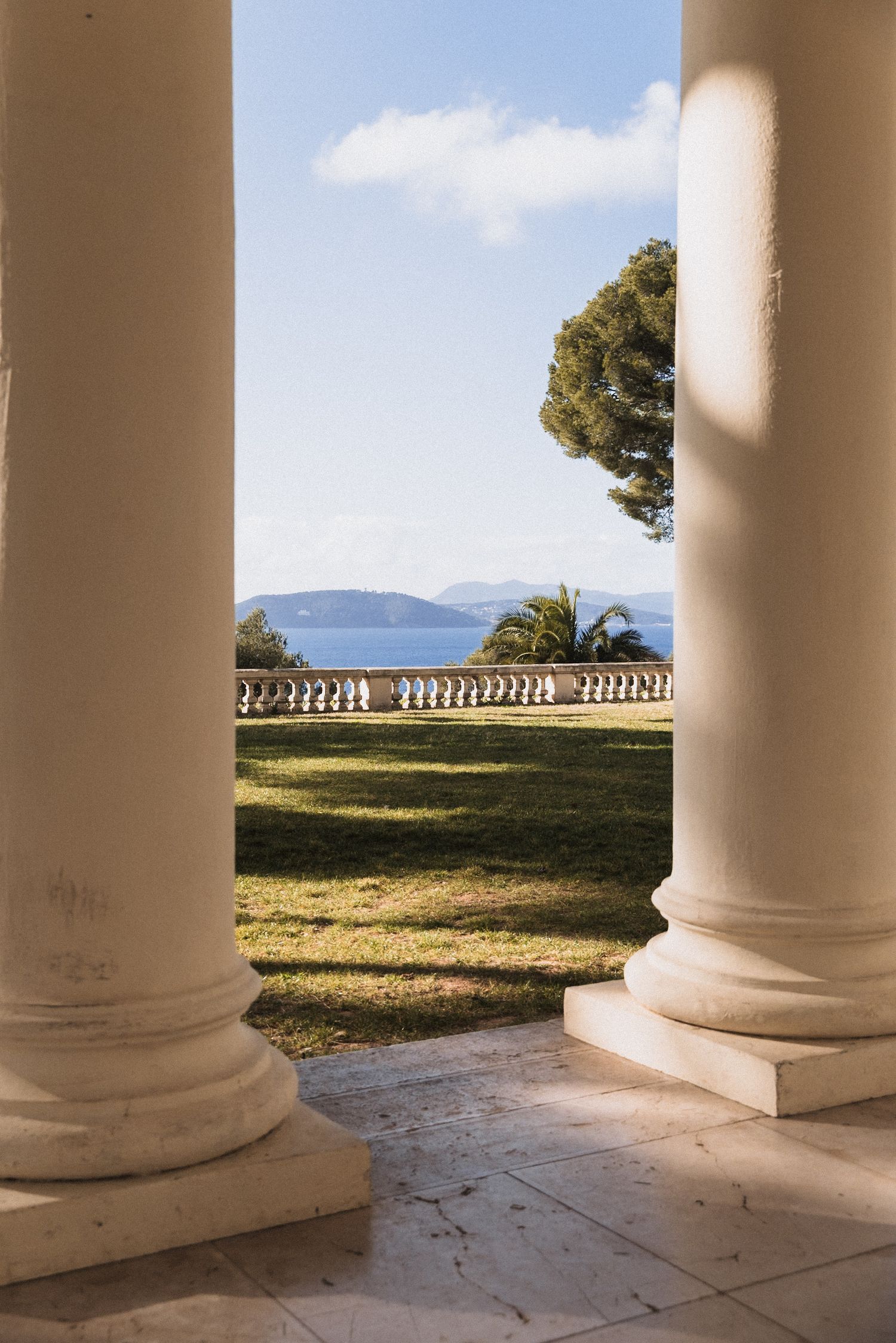 White stone columns frame a scenic coastal view with distant mountains and a decorative balustrade railing visible.