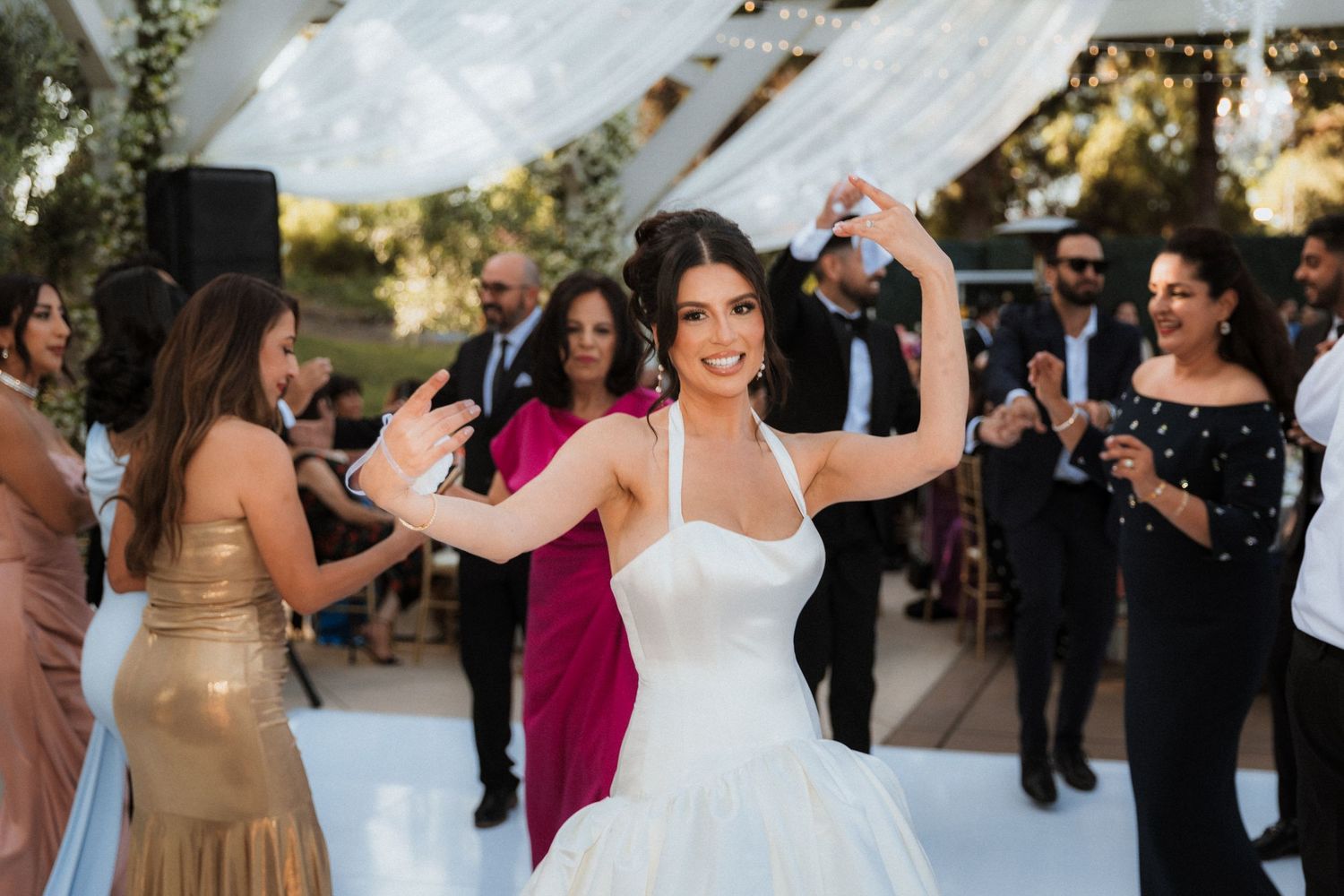 Bride in white ball gown dancing joyfully at outdoor wedding reception with guests celebrating around her.
