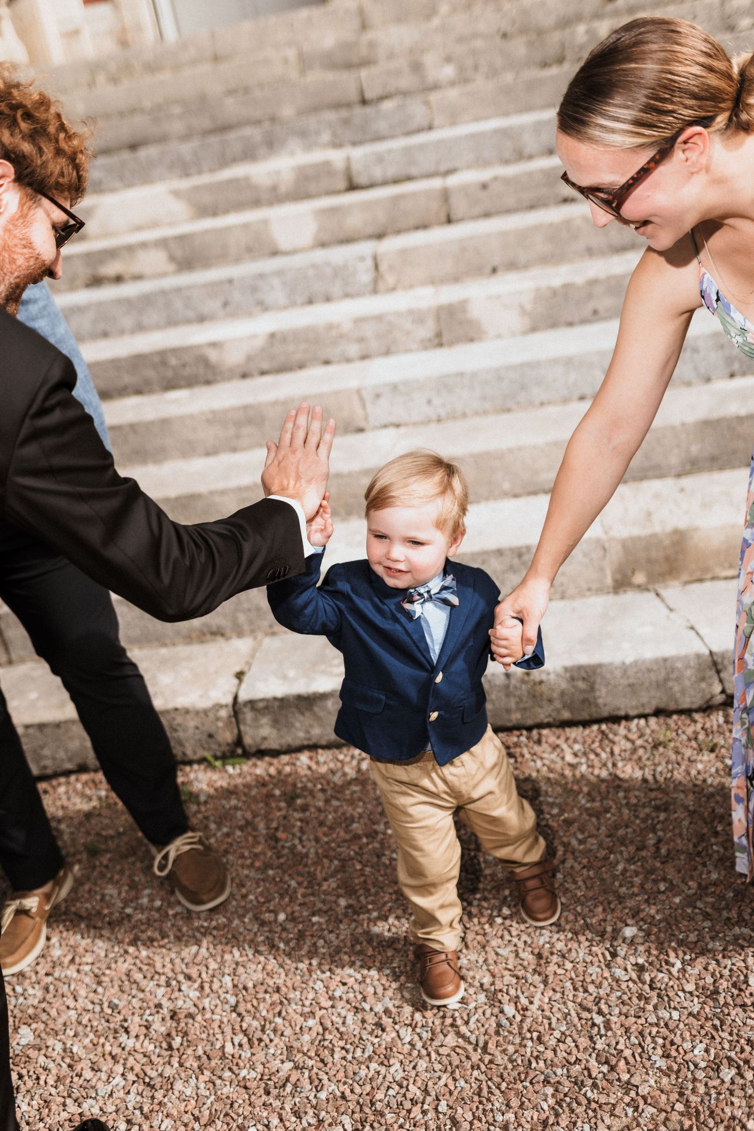 A toddler in a navy blazer and khakis gives a high five while walking up stone steps.
