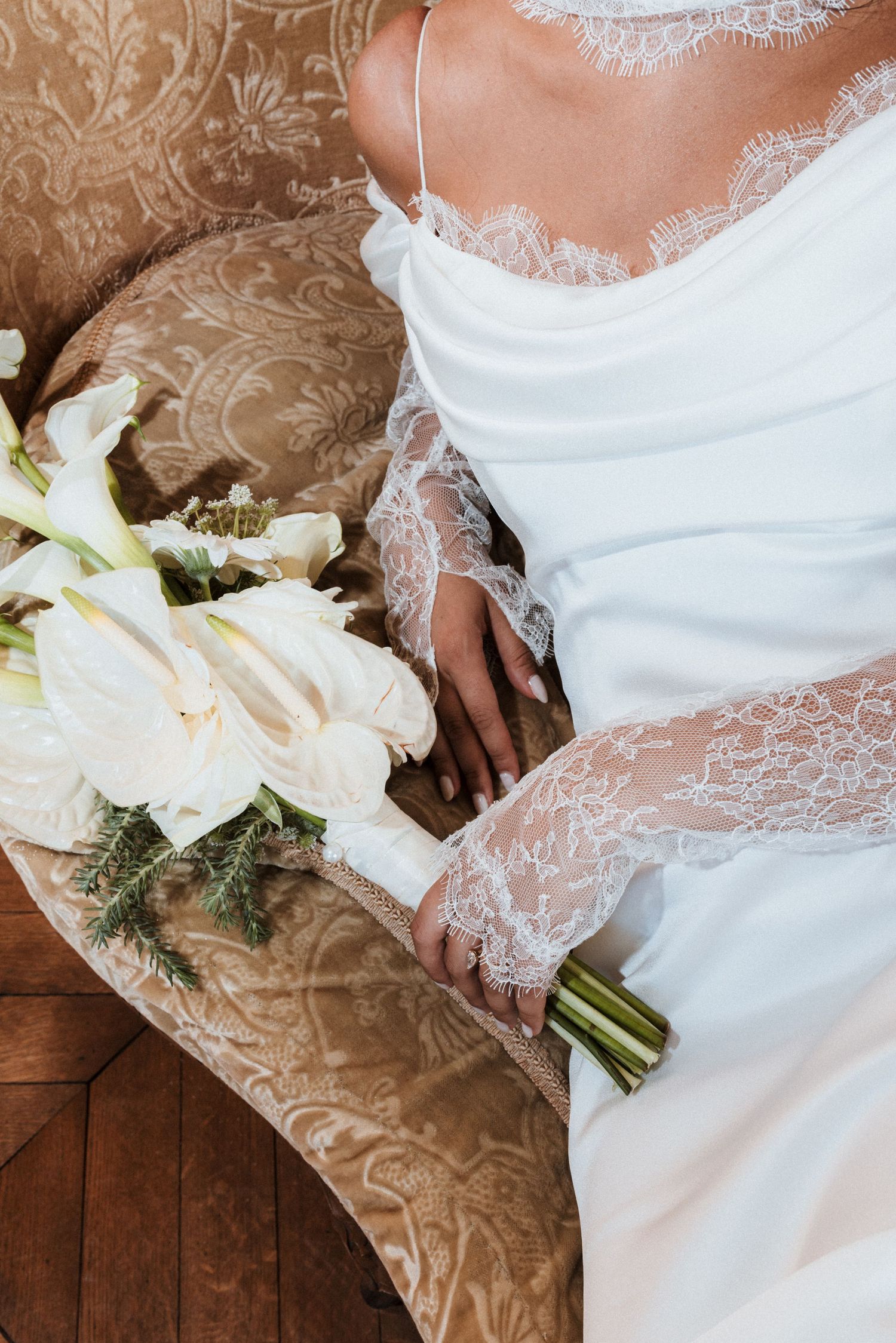 Close-up details of a white lace wedding dress with delicate floral bouquet against a wooden floor background.