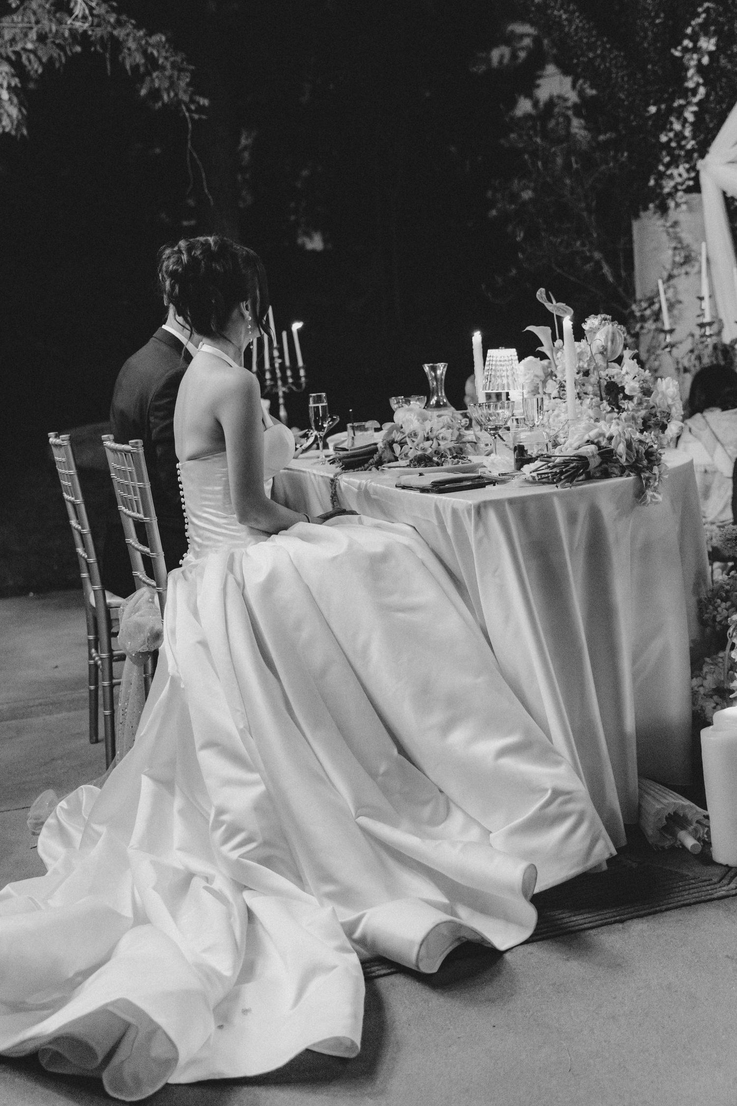 Black and white photo of a bride in a elegant gown seated at a decorated wedding reception table with candles.