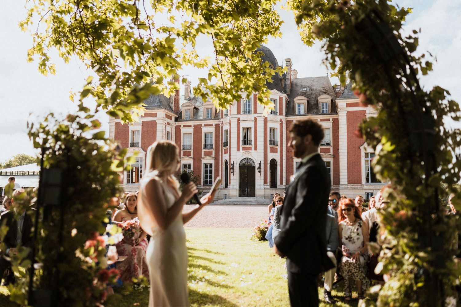 Outdoor wedding ceremony at a grand French château, bride and groom exchange vows under a lush green floral arch.