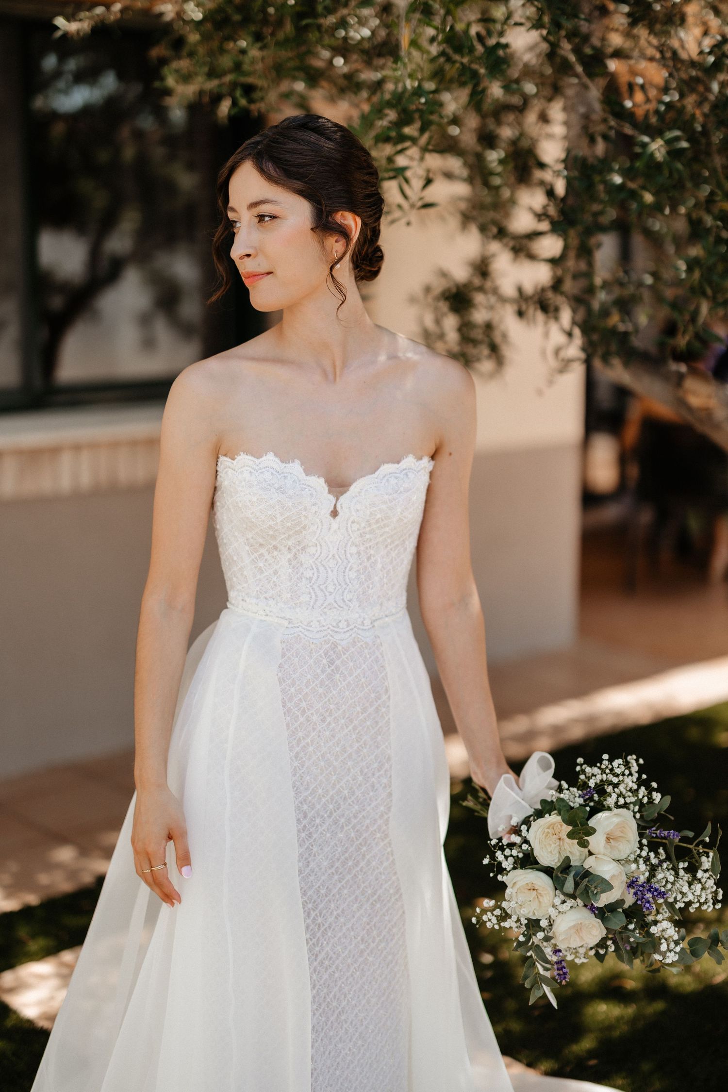 Elegant white wedding dress with lace bodice and flowing tulle skirt photographed in natural sunlight with floral bouquet. Les Moles.