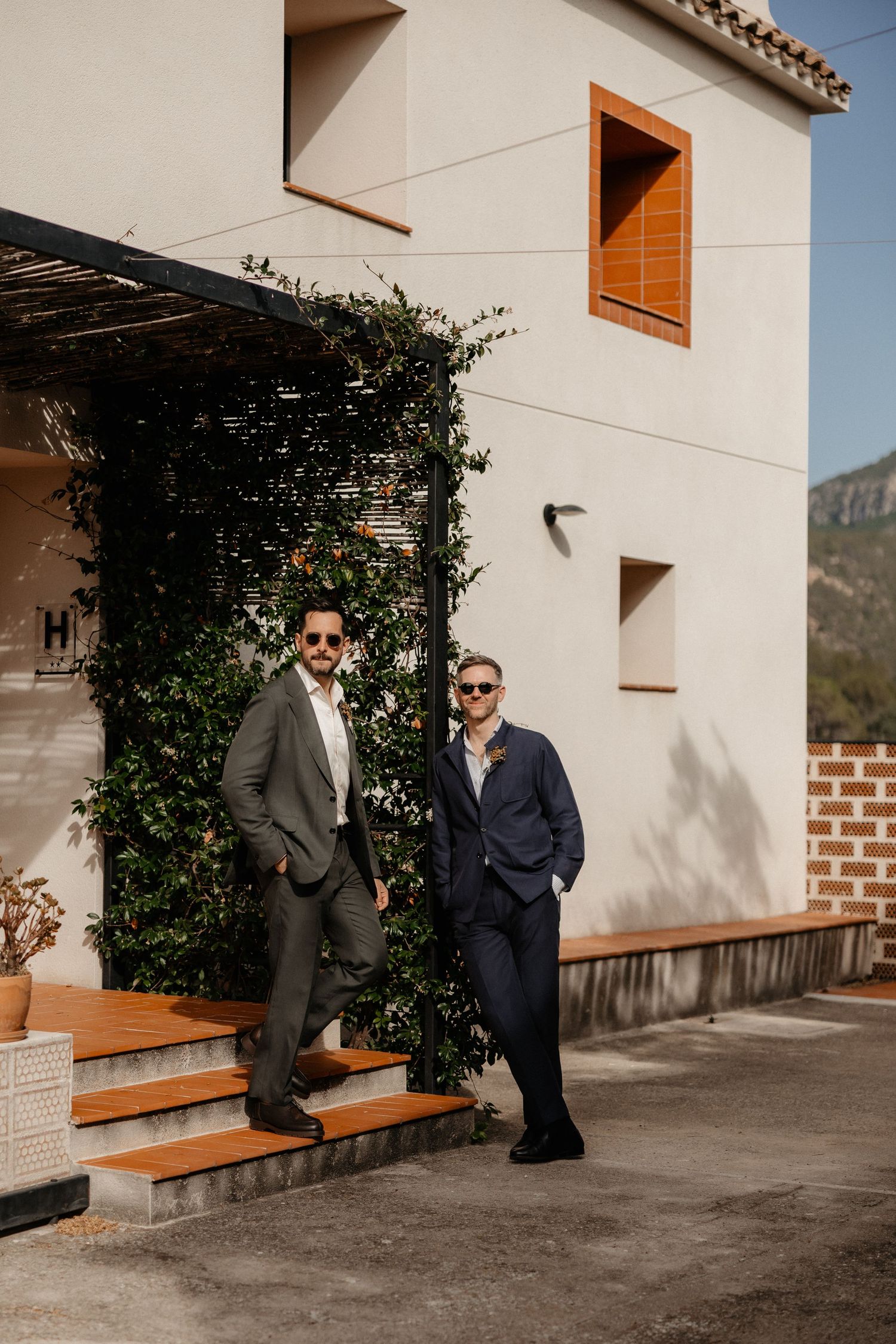 Two men in suits stand against a white building wall with climbing vines and wooden benches in warm sunlight. Wedding in Mas de l'Illa.