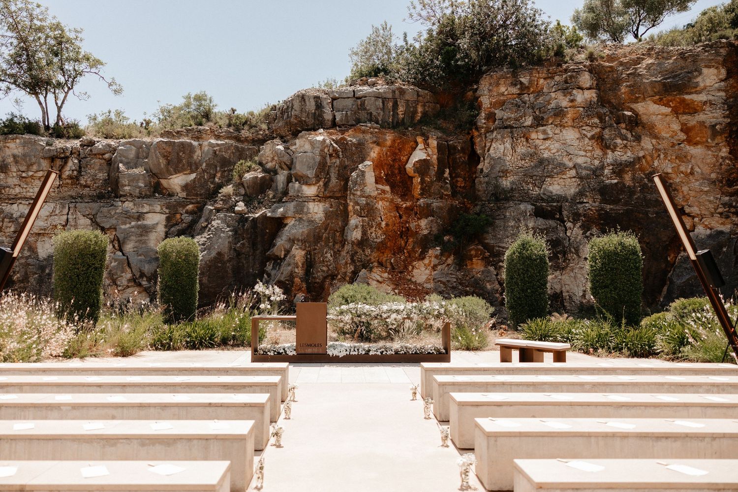 Stone benches arranged in rows face a rustic rock wall altar with stairs in an outdoor garden setting. Wedding in Les Moles.