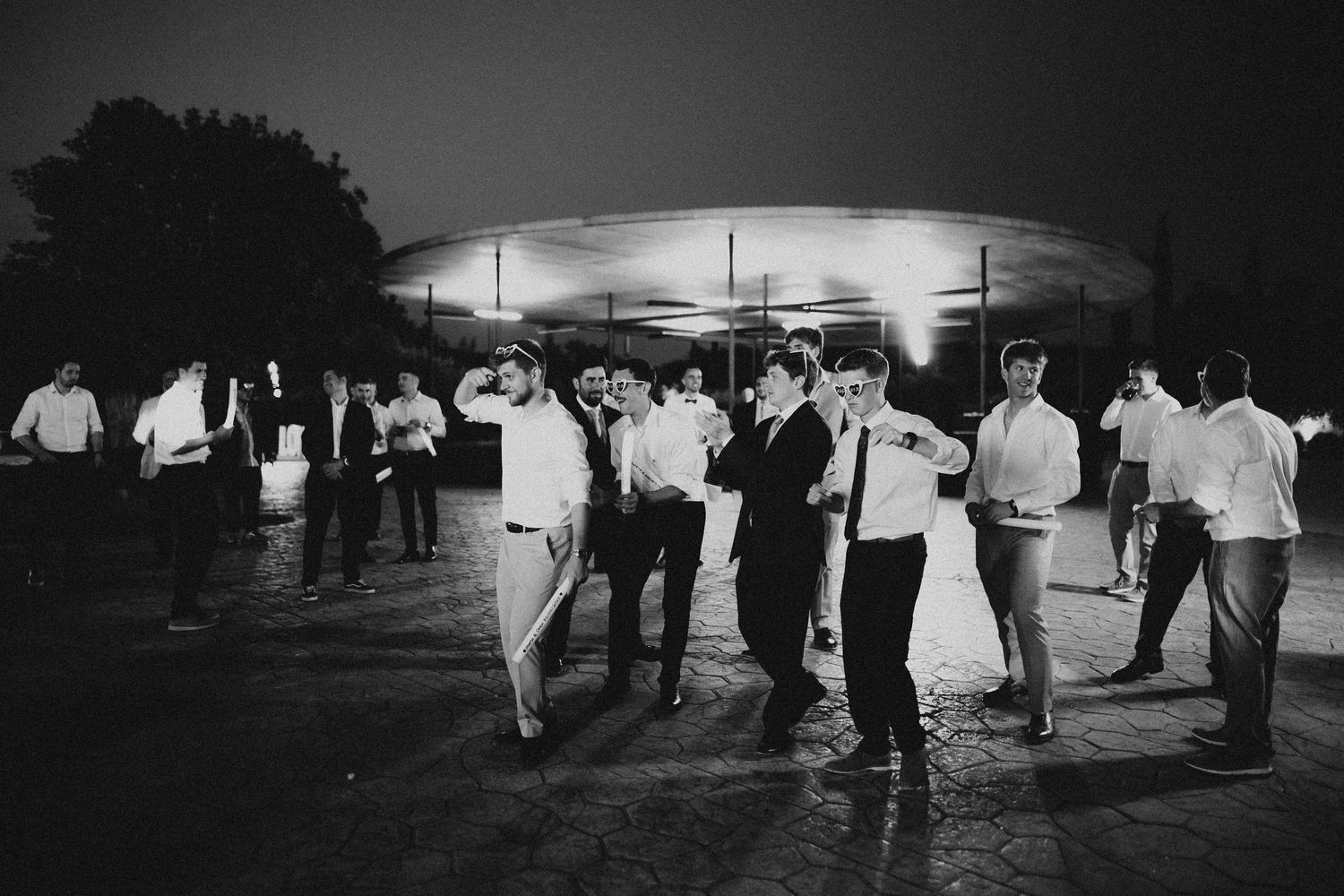 Black and white photo of people dancing outdoors at night under illuminated venue lighting. Wedding in Les Moles.