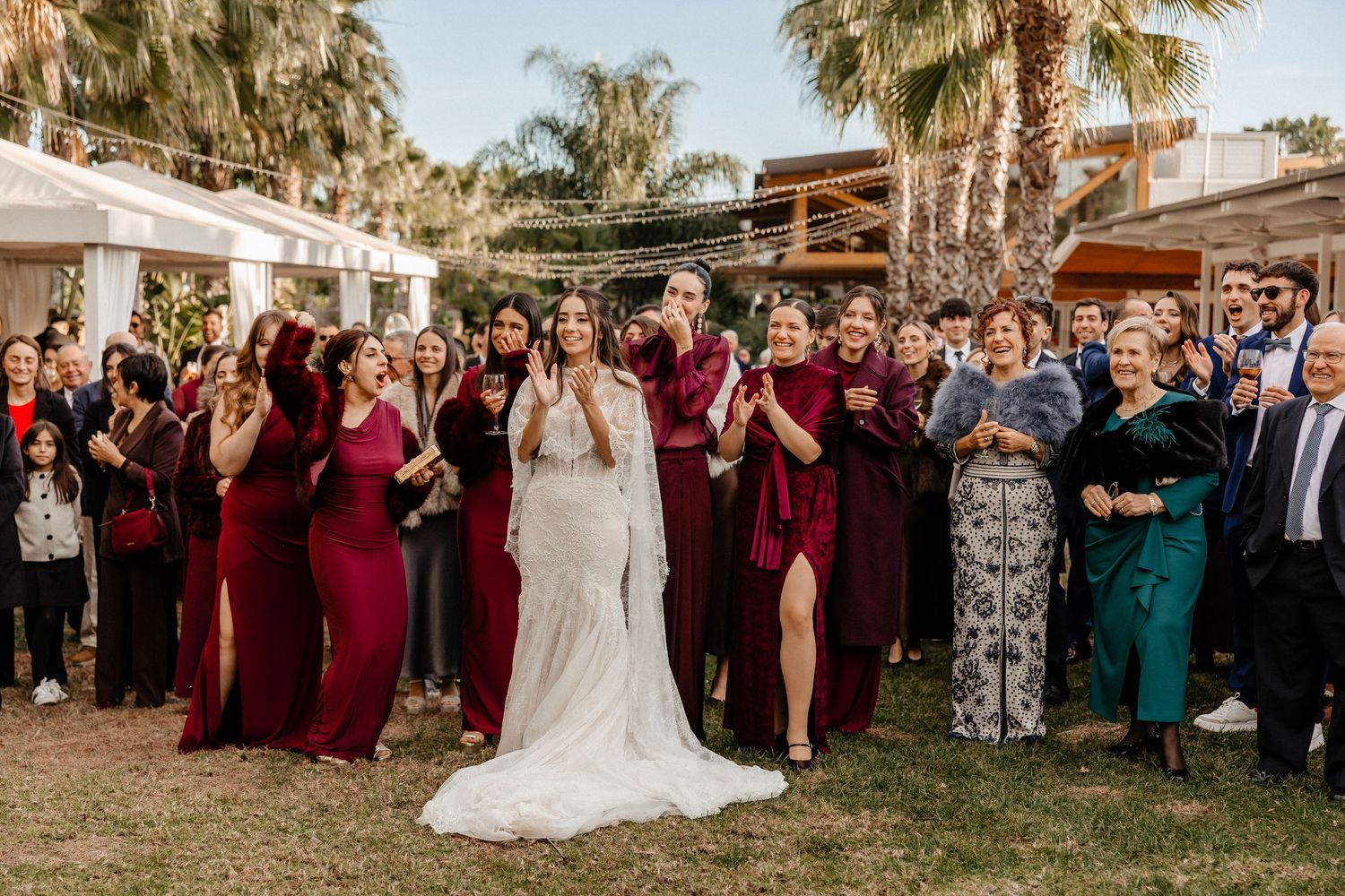 Wedding party and guests gather outdoors at sunset under palm trees for group photos at a tropical venue. Wedding in Jardins del Tancat.