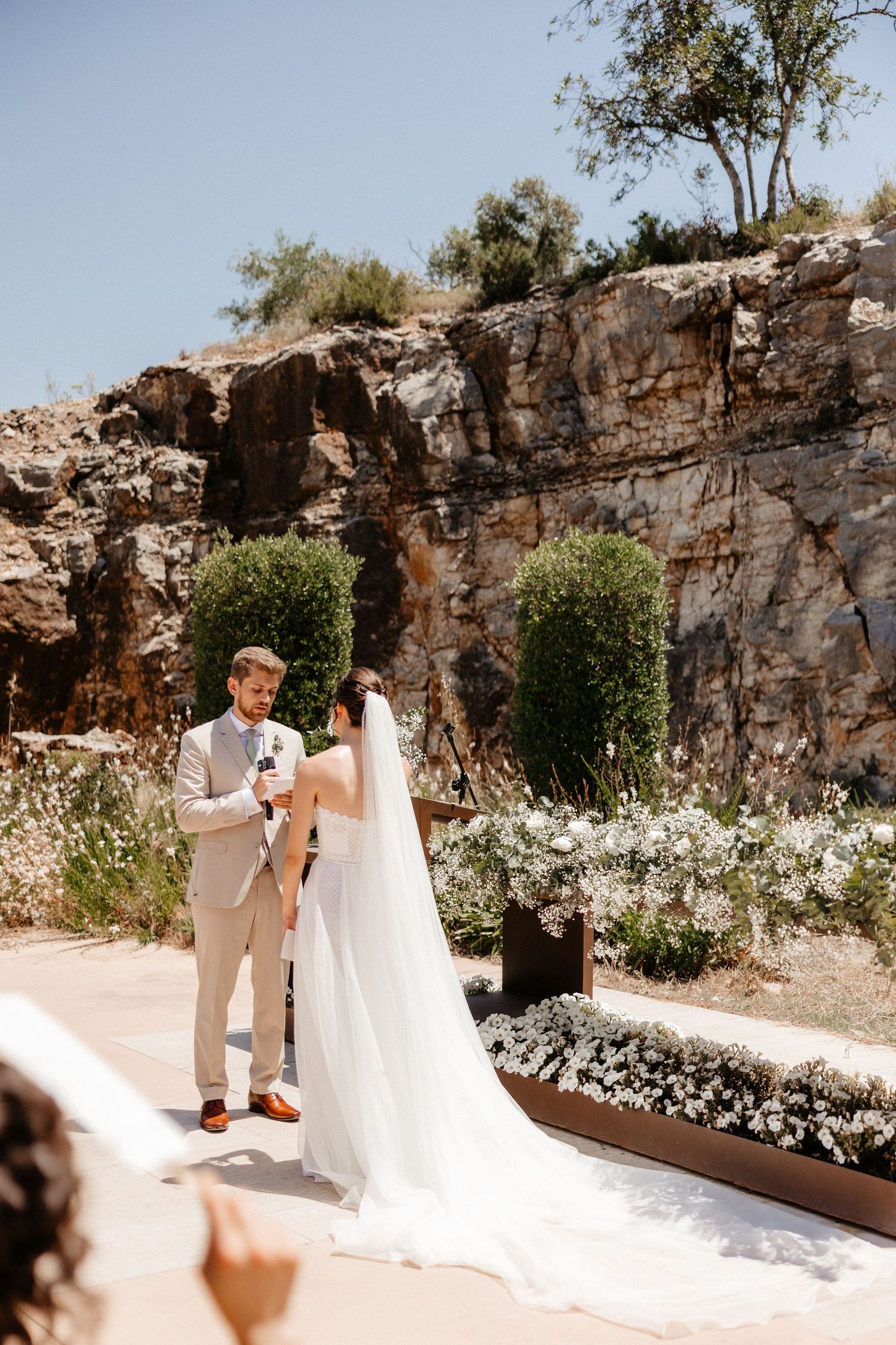 A couple shares a romantic moment against a rustic stone wall backdrop adorned with delicate white flowers. Wedding in Les Moles.