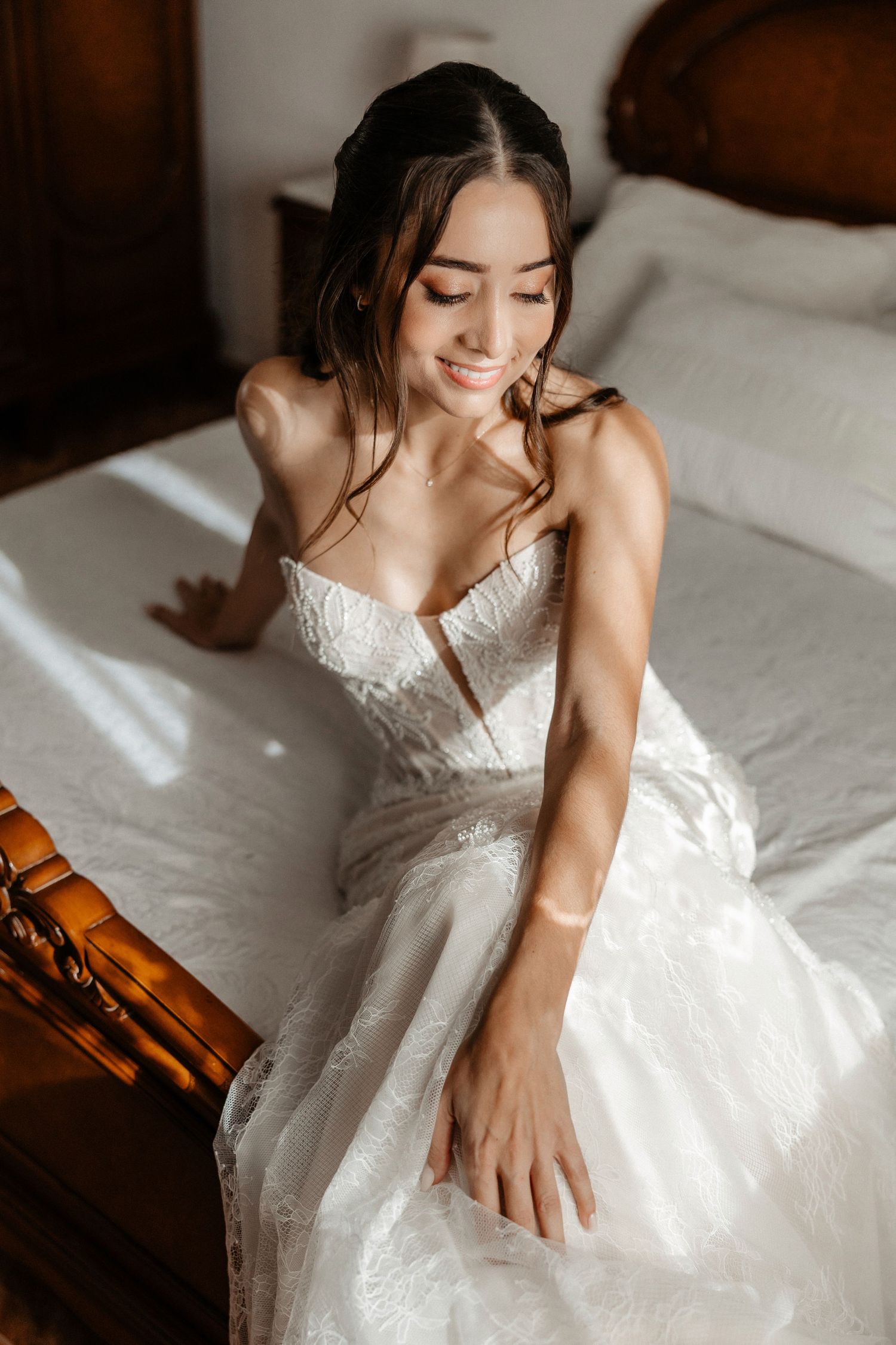 A bride in a white lace wedding dress sits gracefully on white bedding in soft natural light. Wedding in Jardins del Tancat.