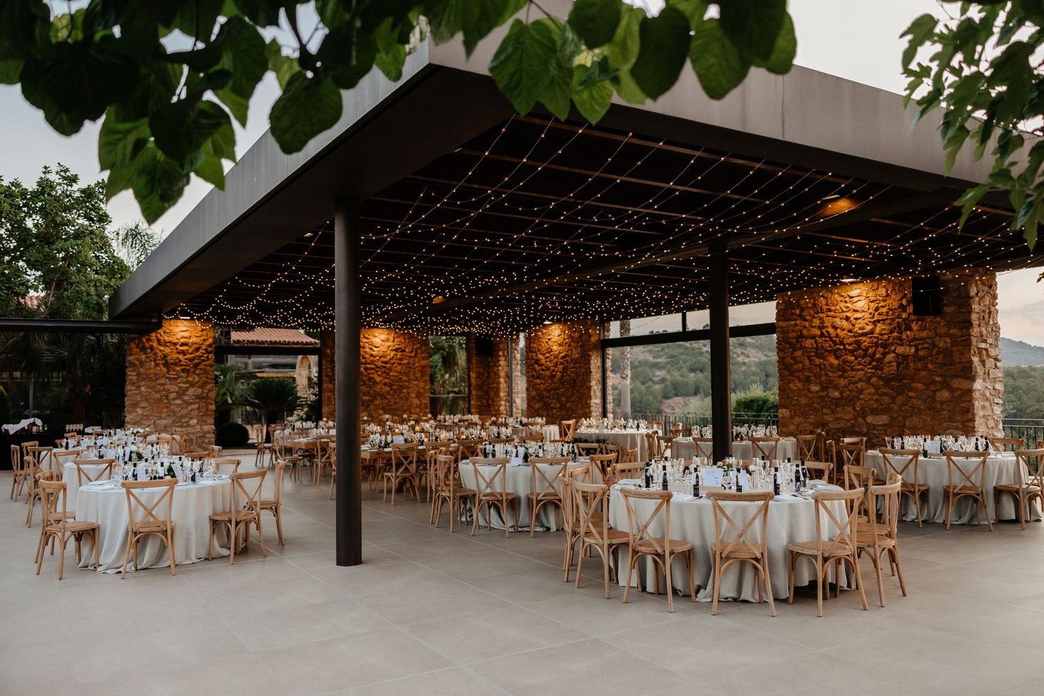 A panoramic view of an elegant rustic wedding reception venue with wooden chairs and exposed brick walls. Masia Les Casotes.