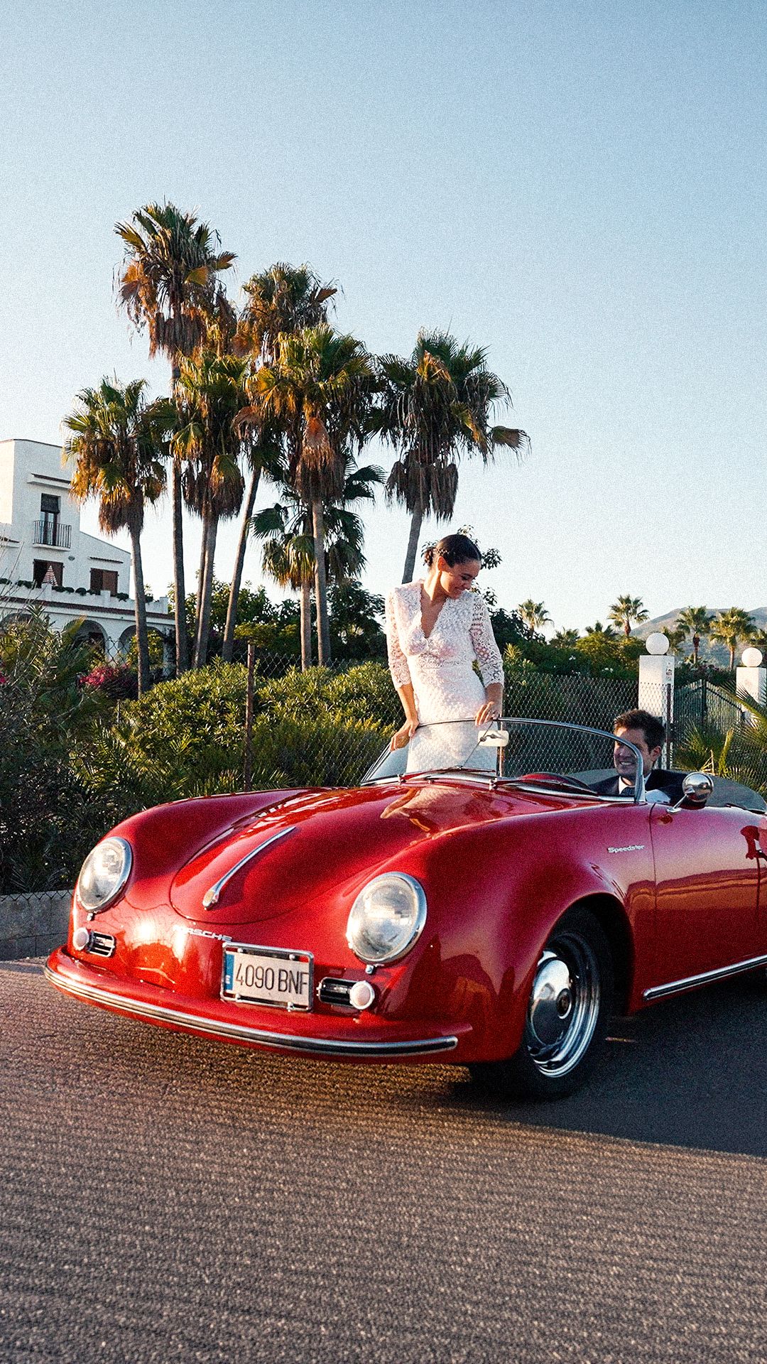 Two people pose with a classic red Porsche 356 convertible near palm trees in a sunny California coastal setting.