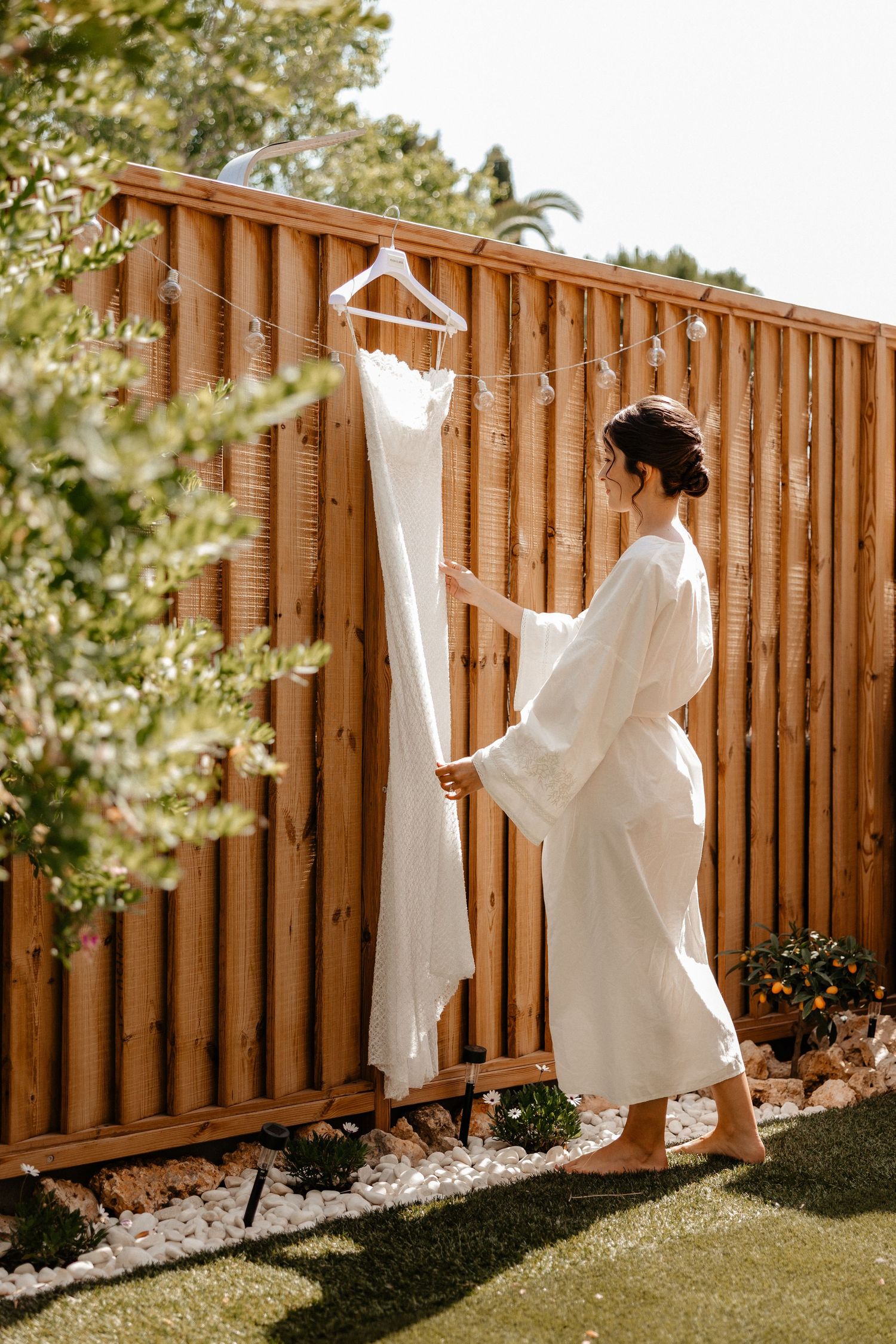 A white wedding dress hanging on a wooden fence in a backyard with sunlit greenery in the background. Wedding in Les Moles.