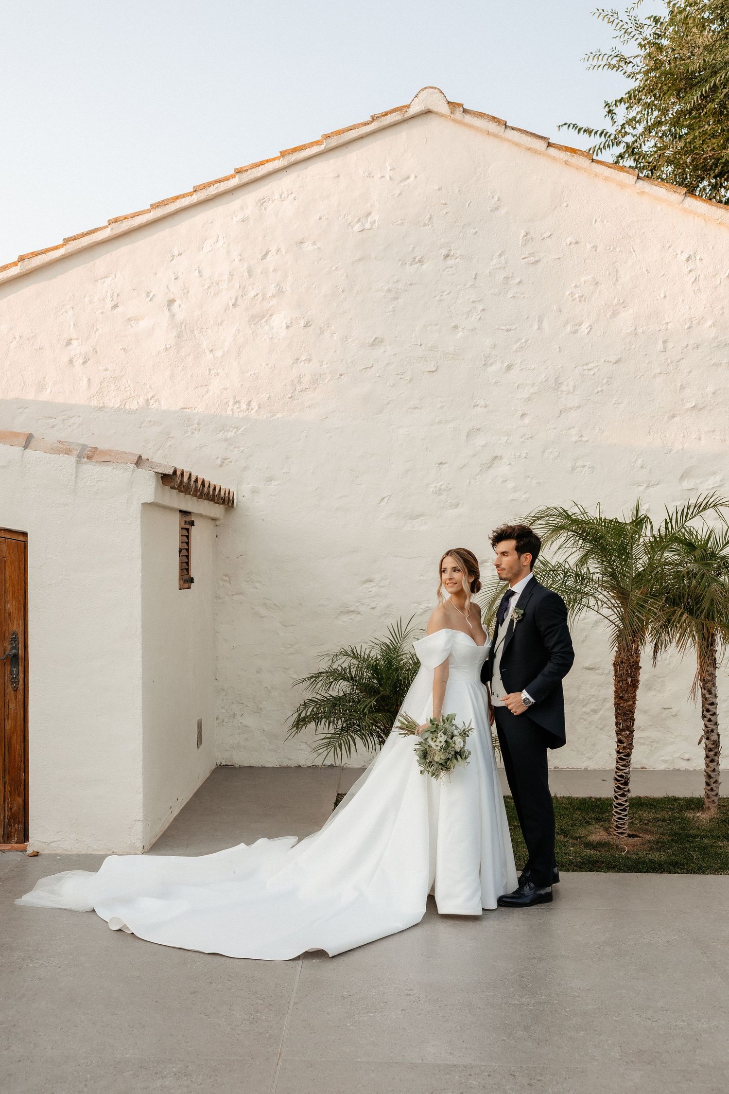 A bride and groom share a romantic moment against a white stucco wall with palm trees in an outdoor courtyard. Masia Les Casotes.