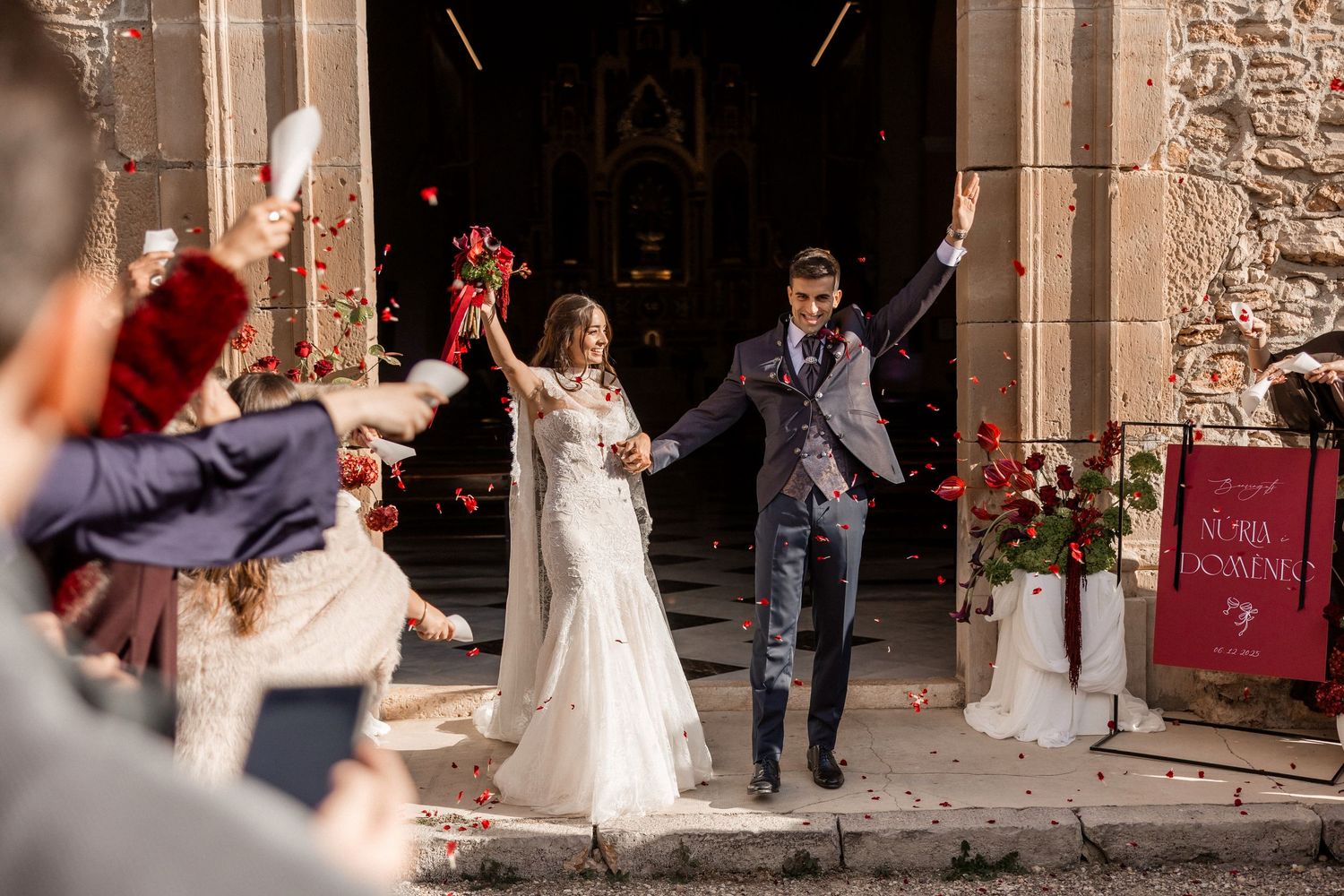 A joyful couple exits a stone church while guests celebrate with confetti and cheers on their wedding day. Wedding in Jardins del Tancat.