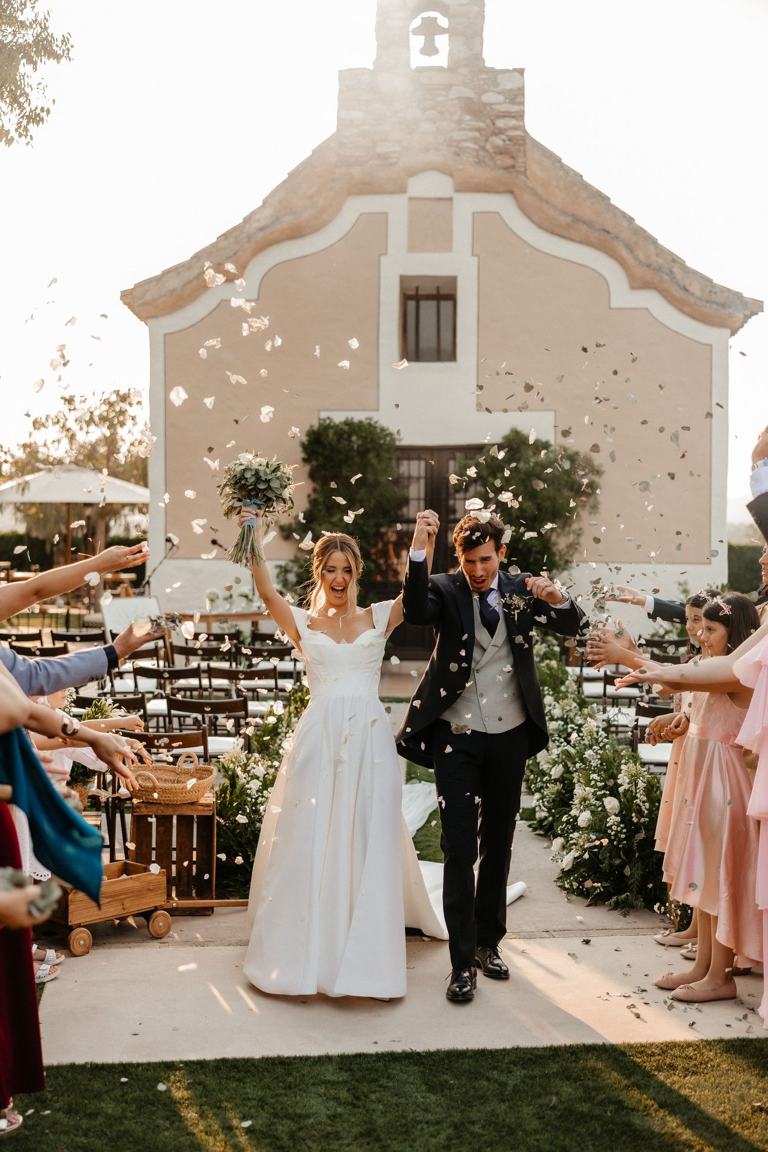 Newlyweds celebrate with raised arms while guests throw confetti outside a charming chapel with pink walls and bell tower. Masia Les Casotes
