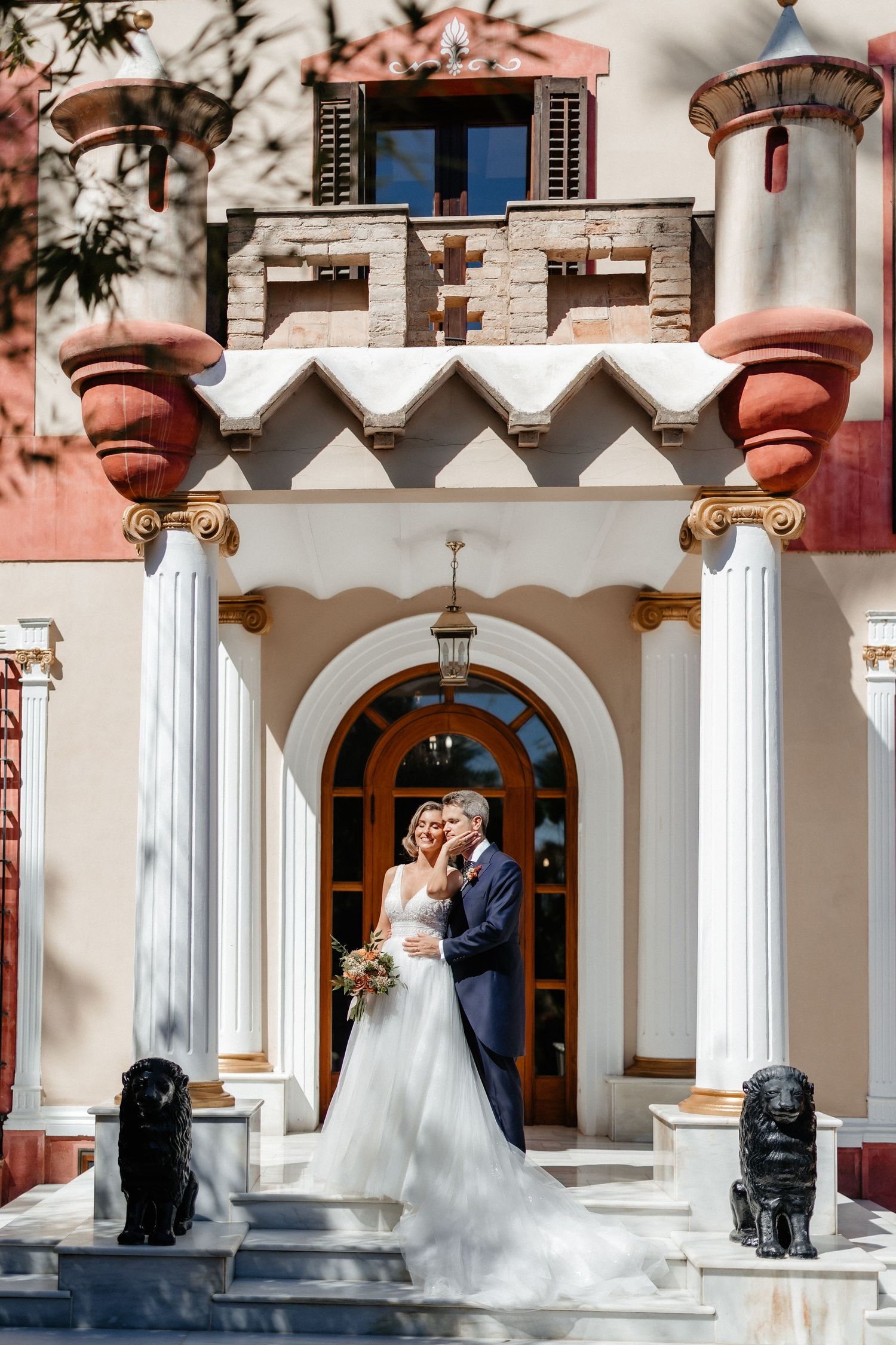 Elegant wedding couple stands in front of ornate white columned entrance with pink and stone architectural details. Wedding in Villa Retiro.