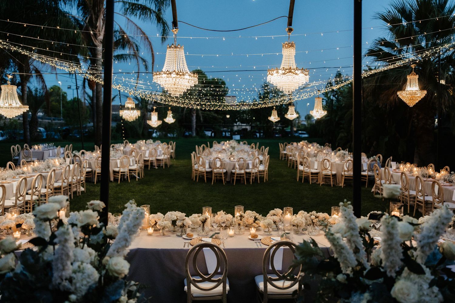 String lights illuminate an elegant outdoor wedding reception setup with white tablecloths and floral centerpieces at dusk. Villa retiro.
