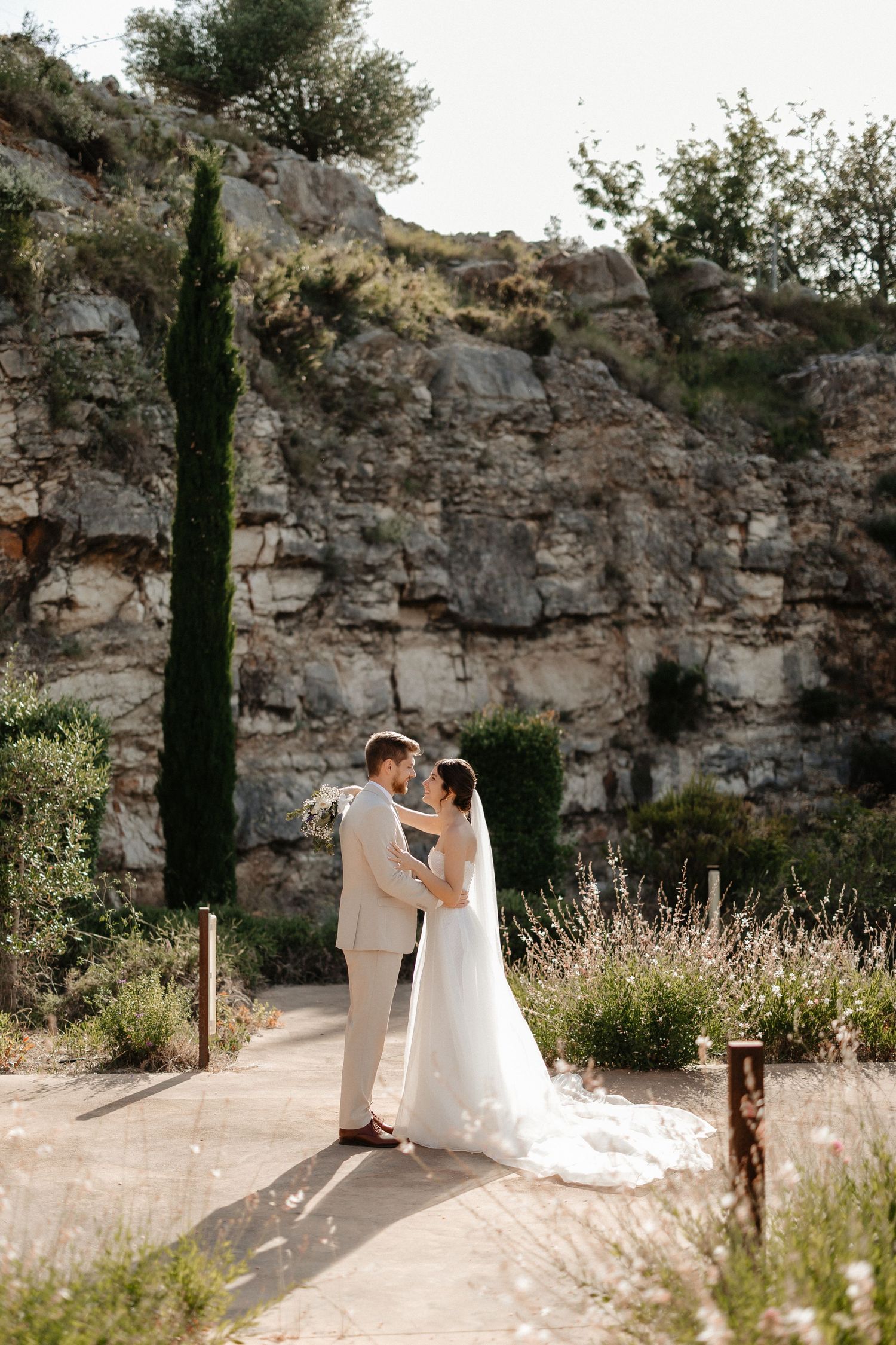 A couple in wedding attire embraces against a rocky cliff backdrop with cypress trees and natural landscaping. Wedding in Les Moles.