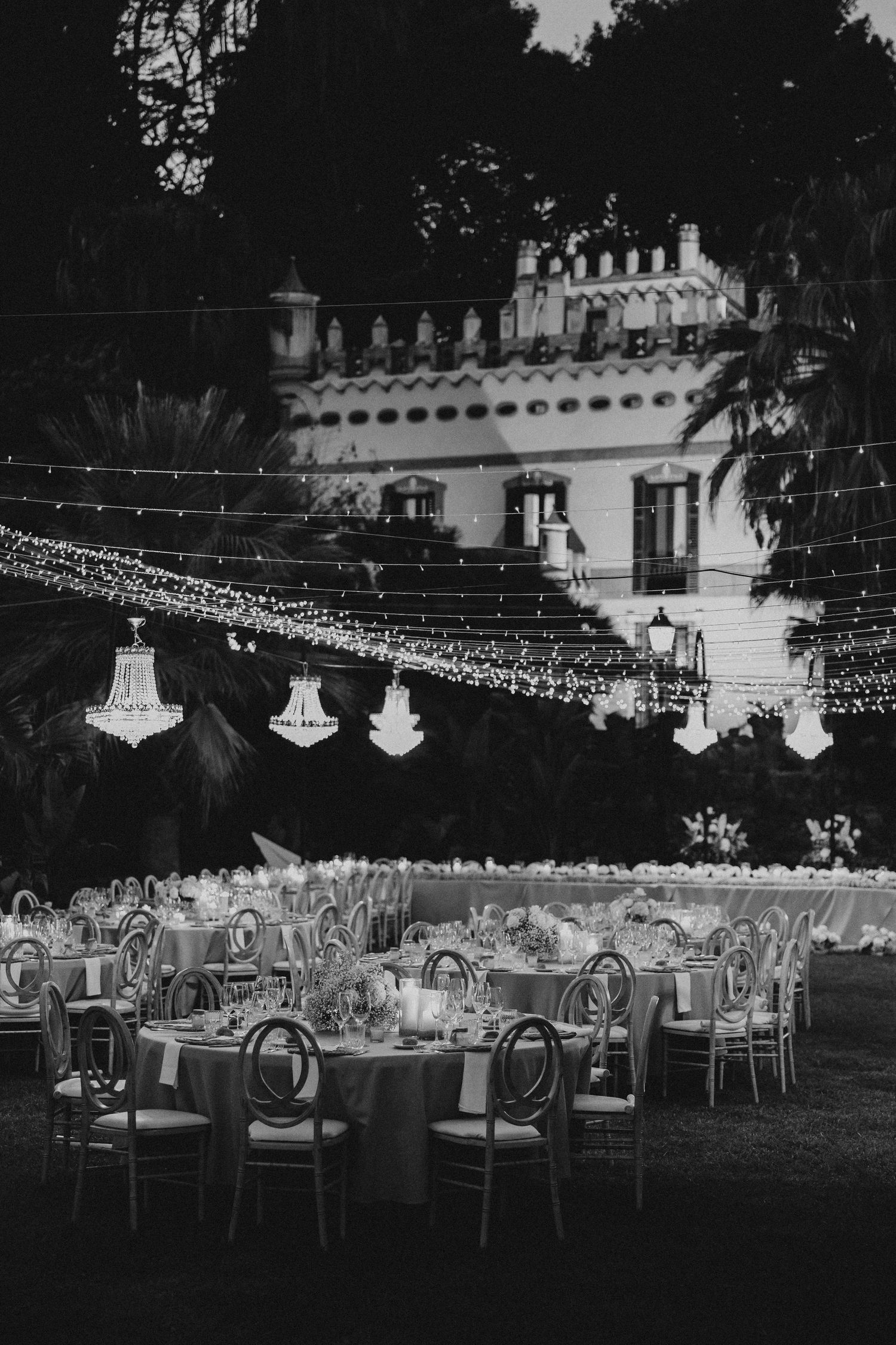 Black and white photo of an elegant outdoor wedding reception with string lights and chandeliers hanging over dining tables. Villa Retiro.