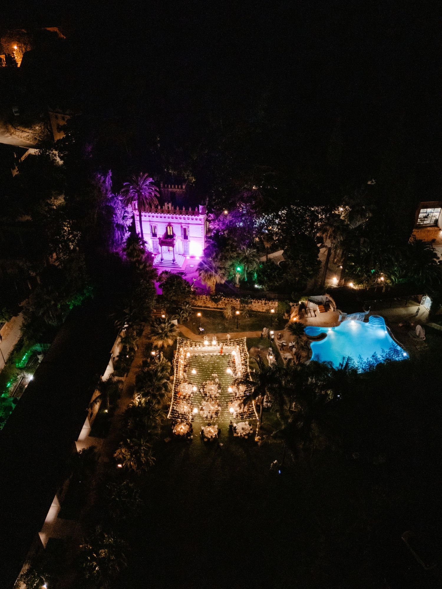 Aerial view of an outdoor wedding reception at night illuminated with colorful lights and decorations in a garden setting. Villa Retiro.