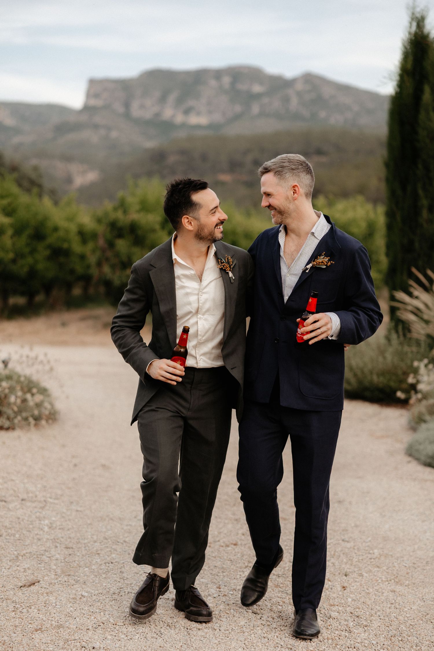 Two people in suits sharing drinks and laughing while walking outdoors with mountains in the background. Mas de l'Illa.