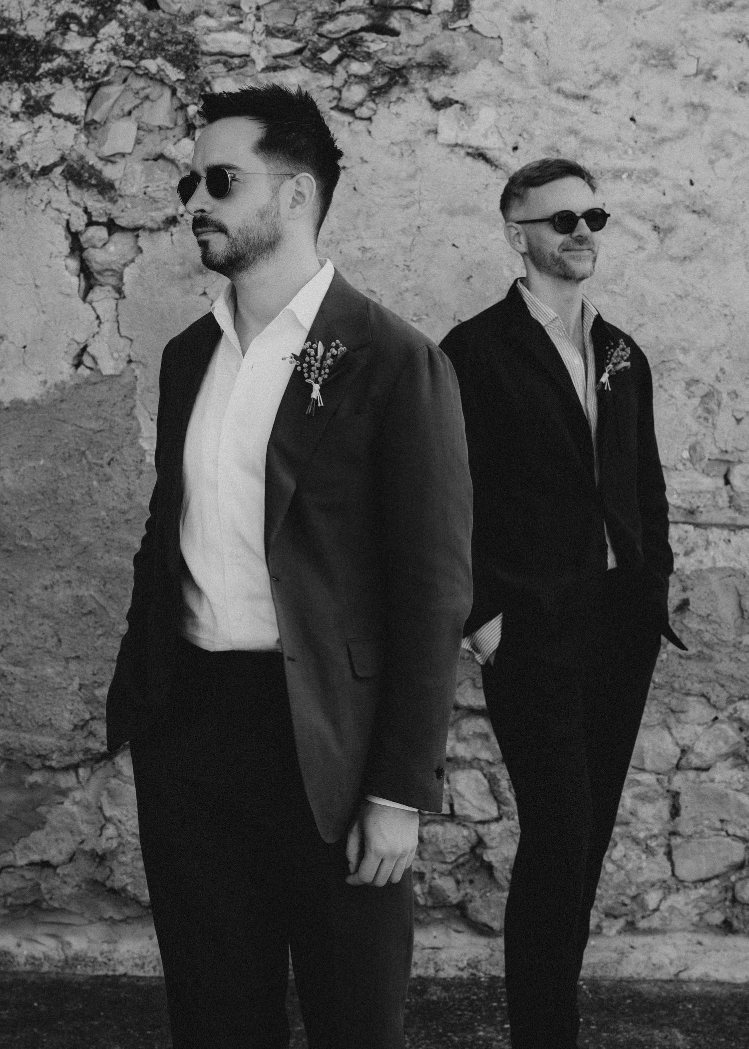 Gay couple in formal attire standing against a stone wall in a black and white photograph.  Wedding in Mas de l'Illa.