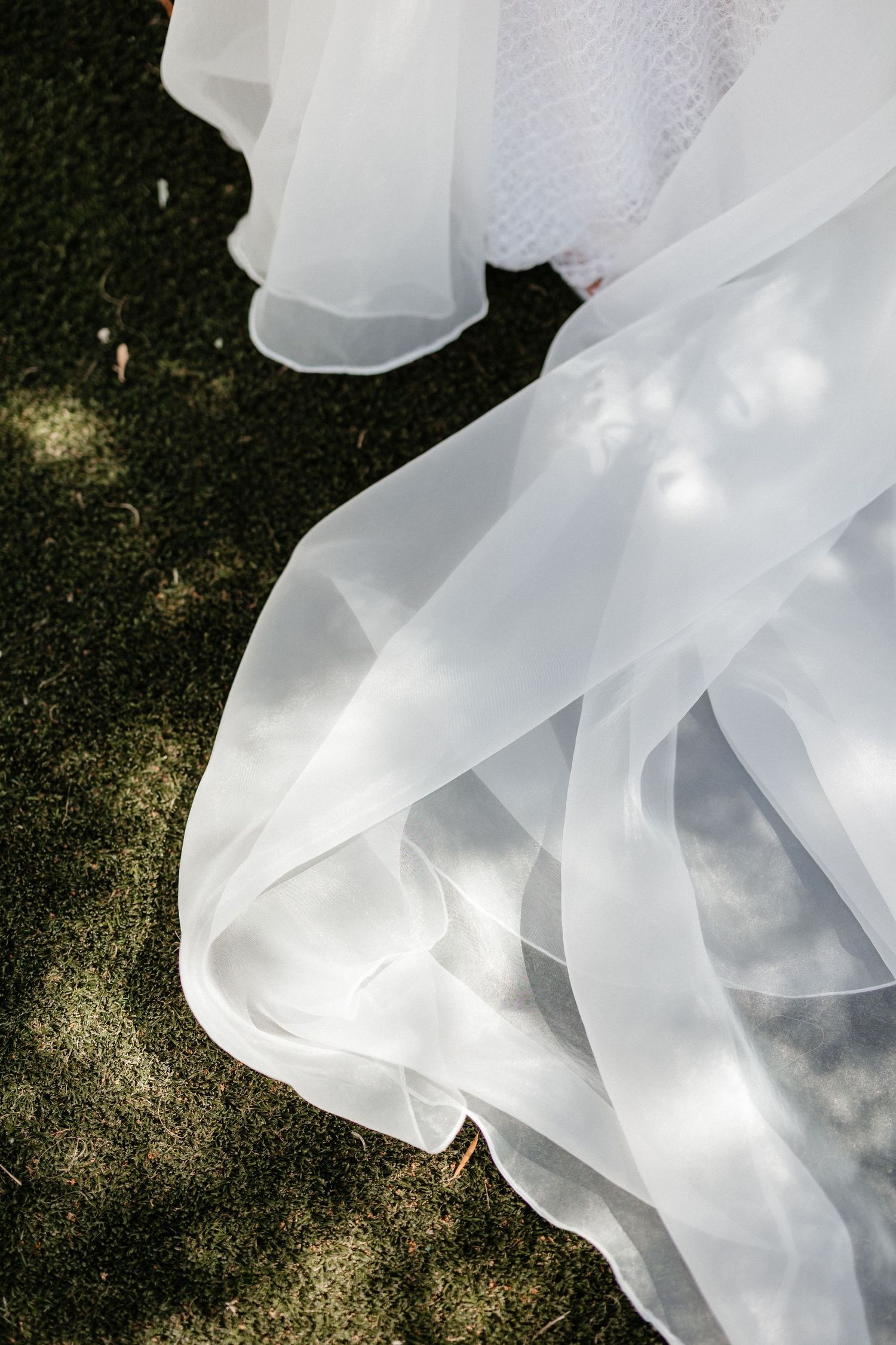 Ethereal white wedding dress fabric flows gracefully against dark grass with dappled sunlight creating a dreamy effect. Wedding in Les Moles