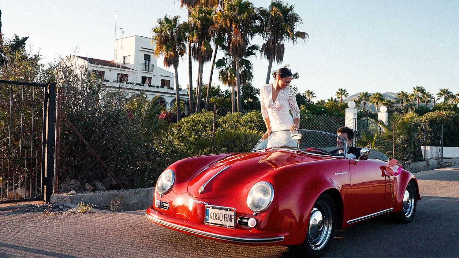 Man standing in red vintage Porsche 356 convertible with palm trees and white building in background.