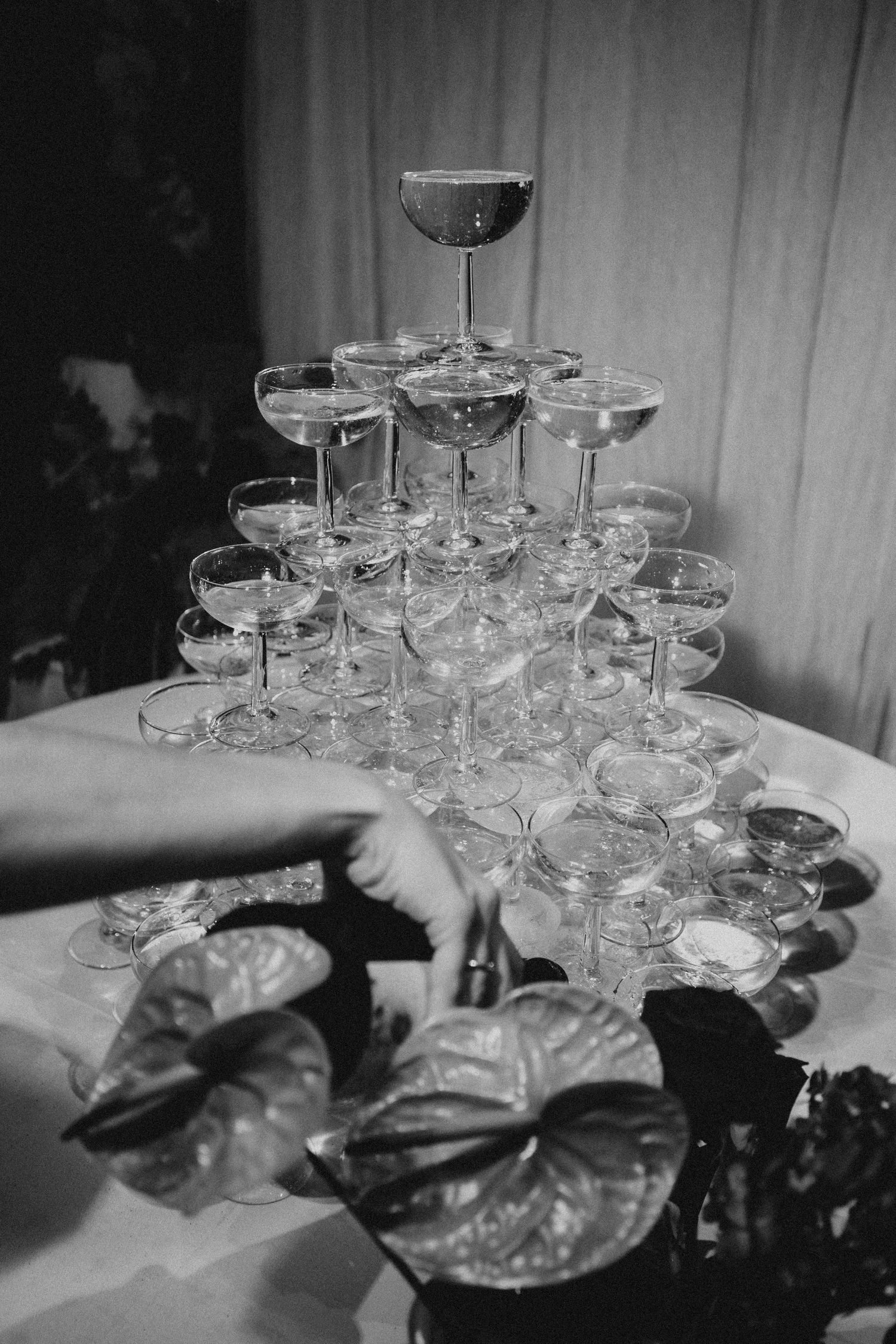 A black and white photograph shows stacked champagne coupes arranged in a tower formation on a table. Wedding in Jardins del Tancat.