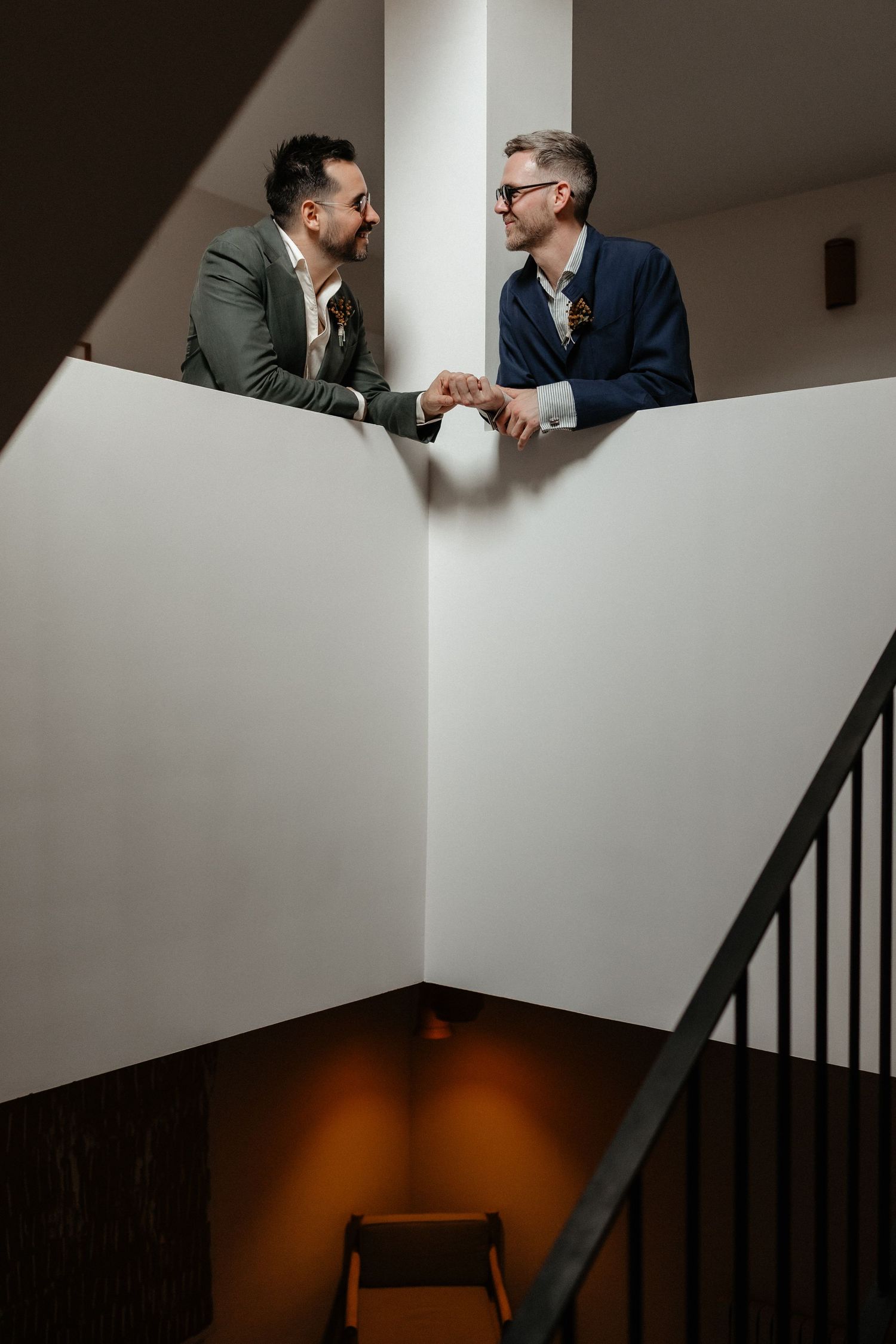Gay couple shake hands and smile while standing on a staircase in a minimalist interior setting. Wedding in Mas de l'Illa.
