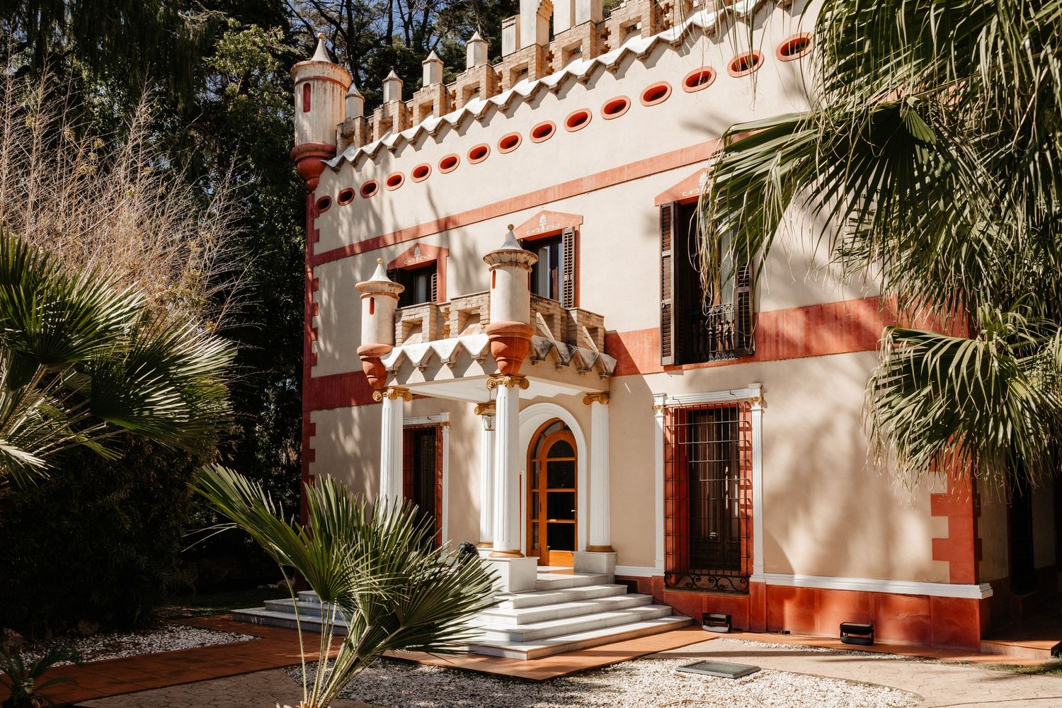 Exterior view of a Mediterranean-style building with red and white striped arches and palm trees in the courtyard. Villa Retiro.