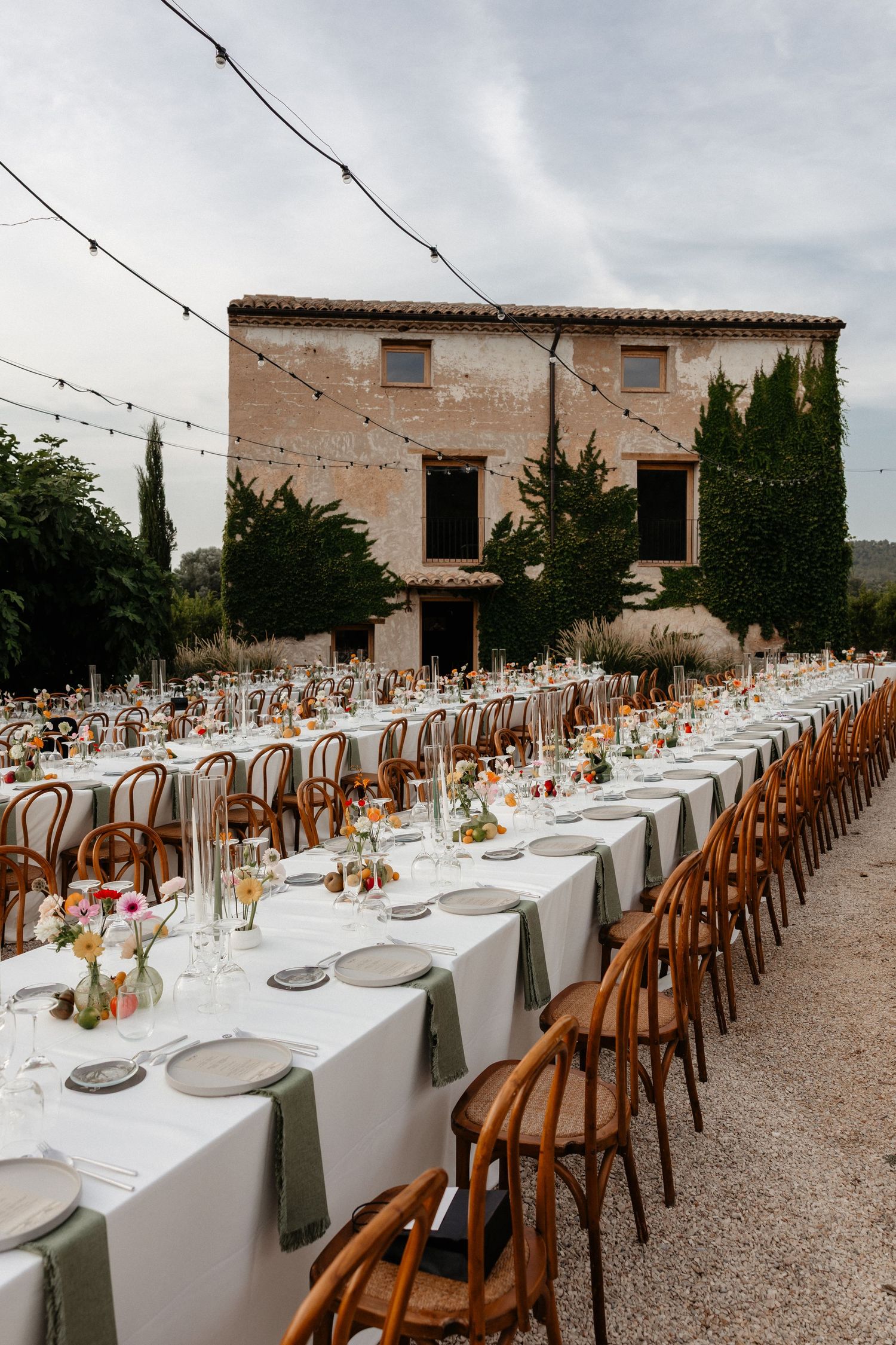 Long outdoor wedding reception tables with white linens and wooden chairs set against rustic stone building backdrop. Mas de l'Ila.